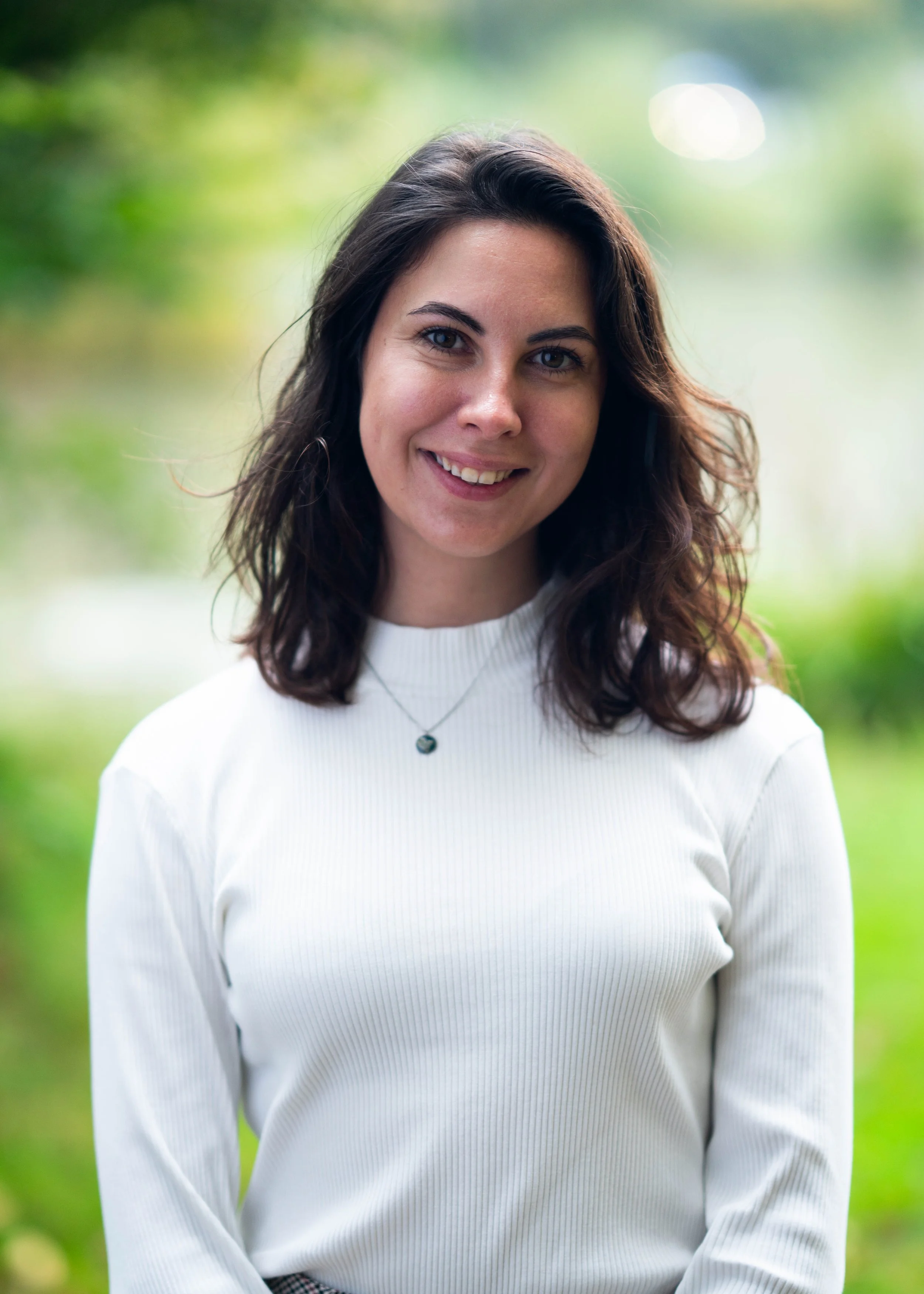 A woman with dark brown, wavy hair and blue eyes smiling, standing outdoors with green blurred background, wearing a white long-sleeve shirt and a small necklace.