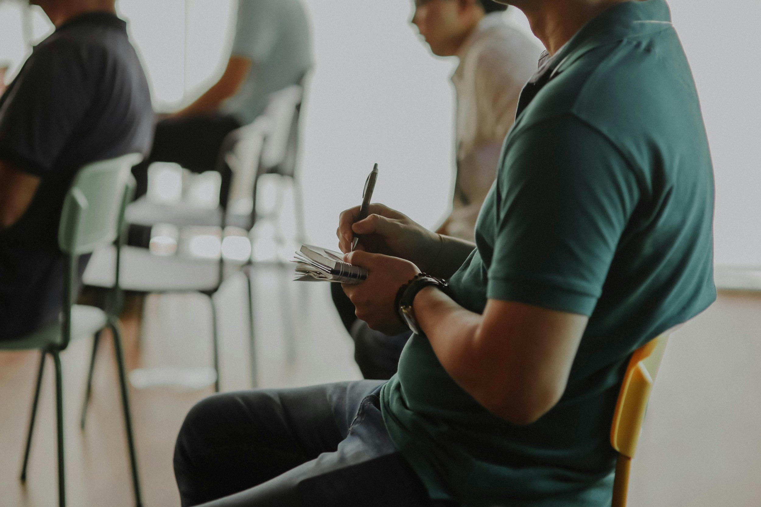Person sitting in a classroom or seminar setting, taking notes in a notepad with a pen.