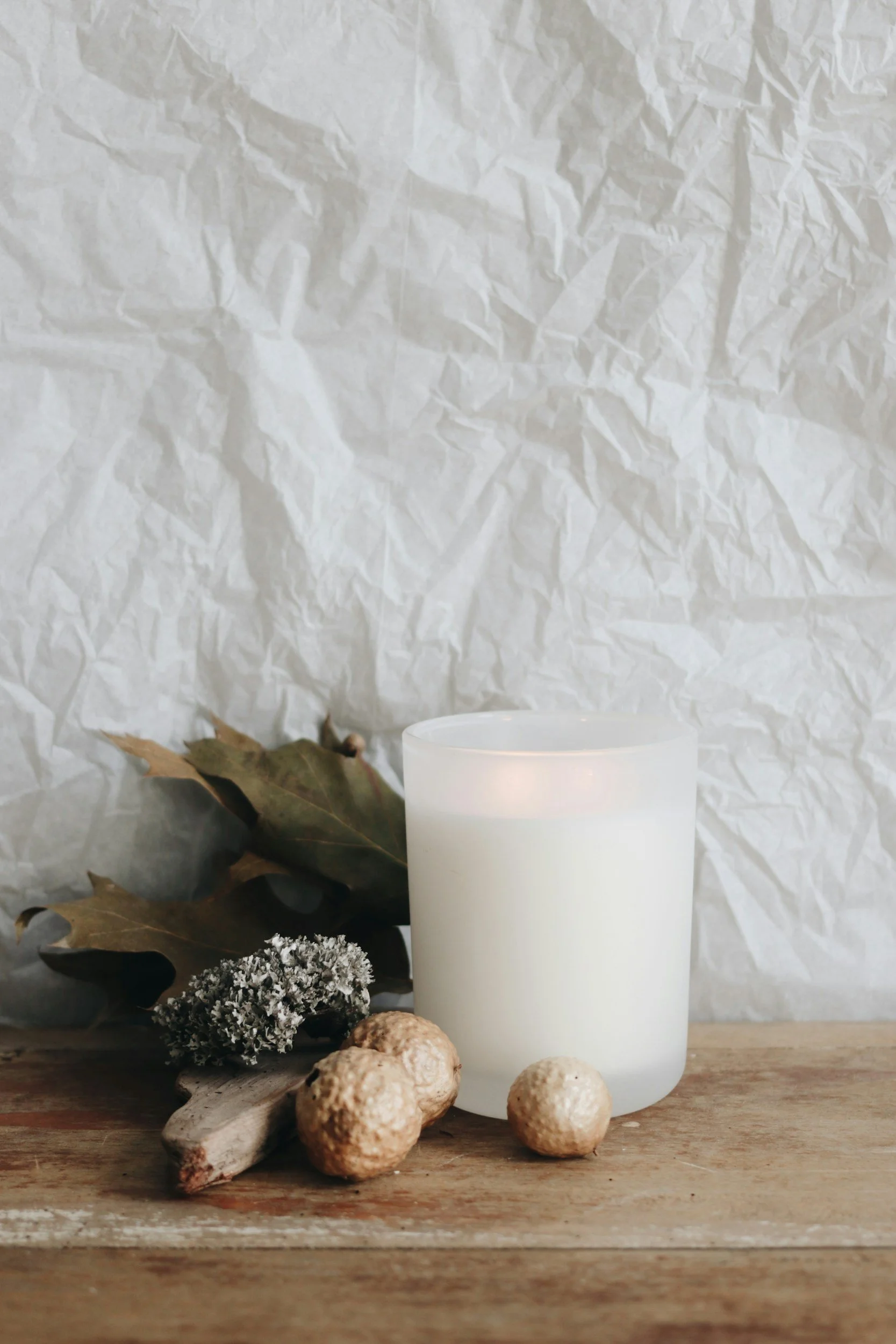 A white candle in a glass holder is on a wooden surface with dried leaves, a piece of driftwood, and round natural stones, against a wrinkled white paper background.
