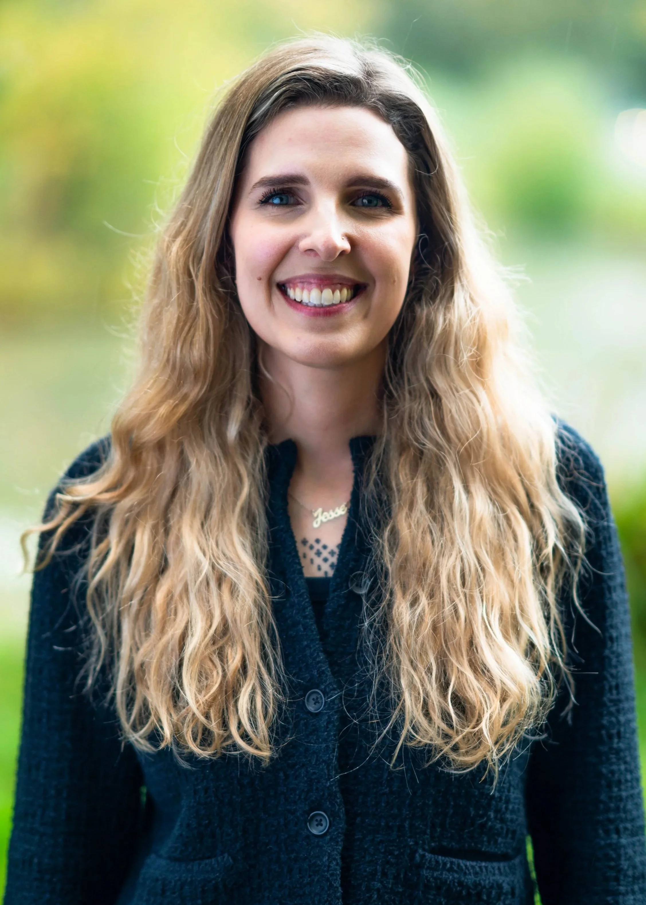 A man with curly dark hair, glasses, and a beard, wearing a dark sweater, sitting indoors with a neutral expression.