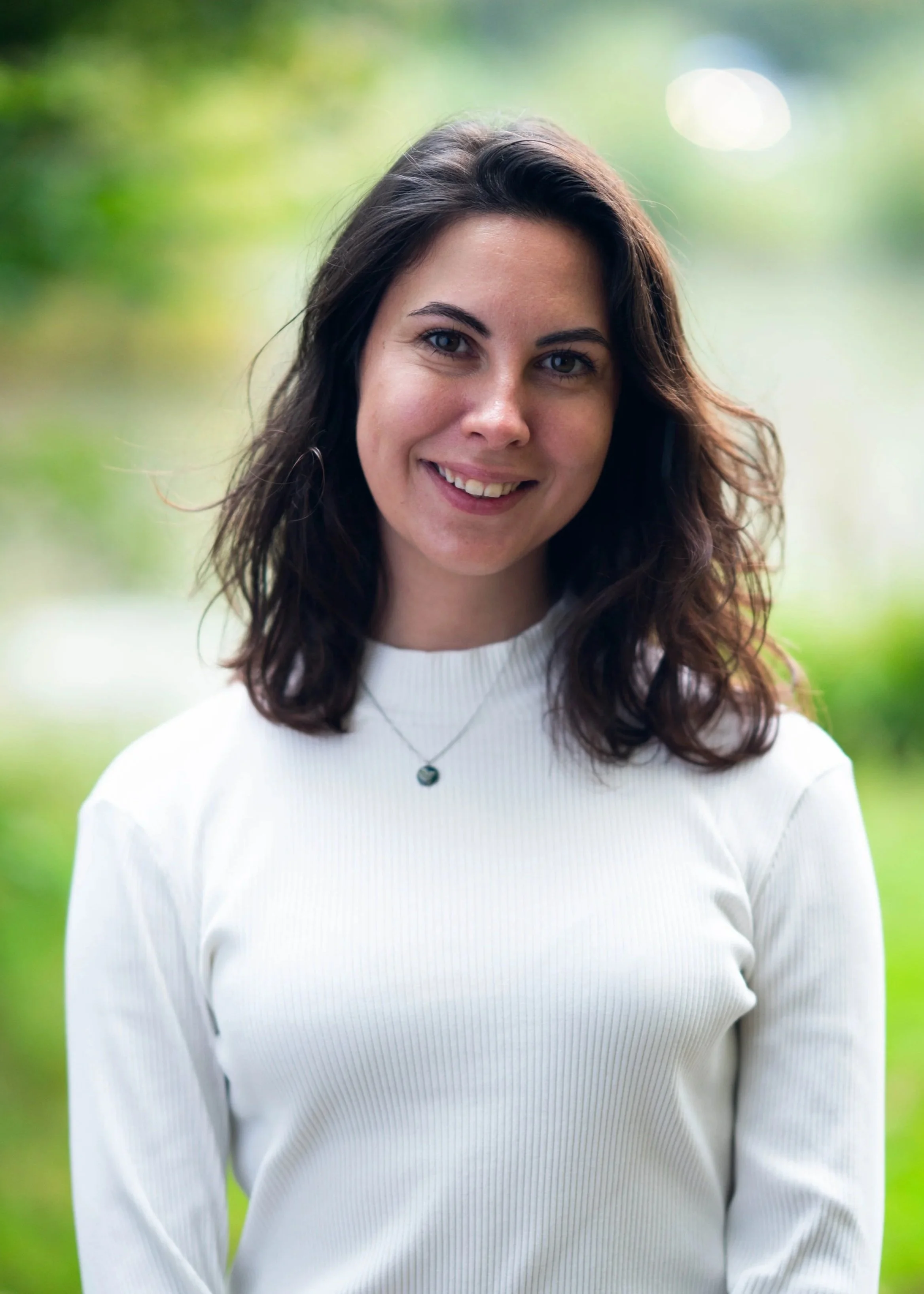 A woman with dark brown hair, wearing a white long sleeve top with trees in the background