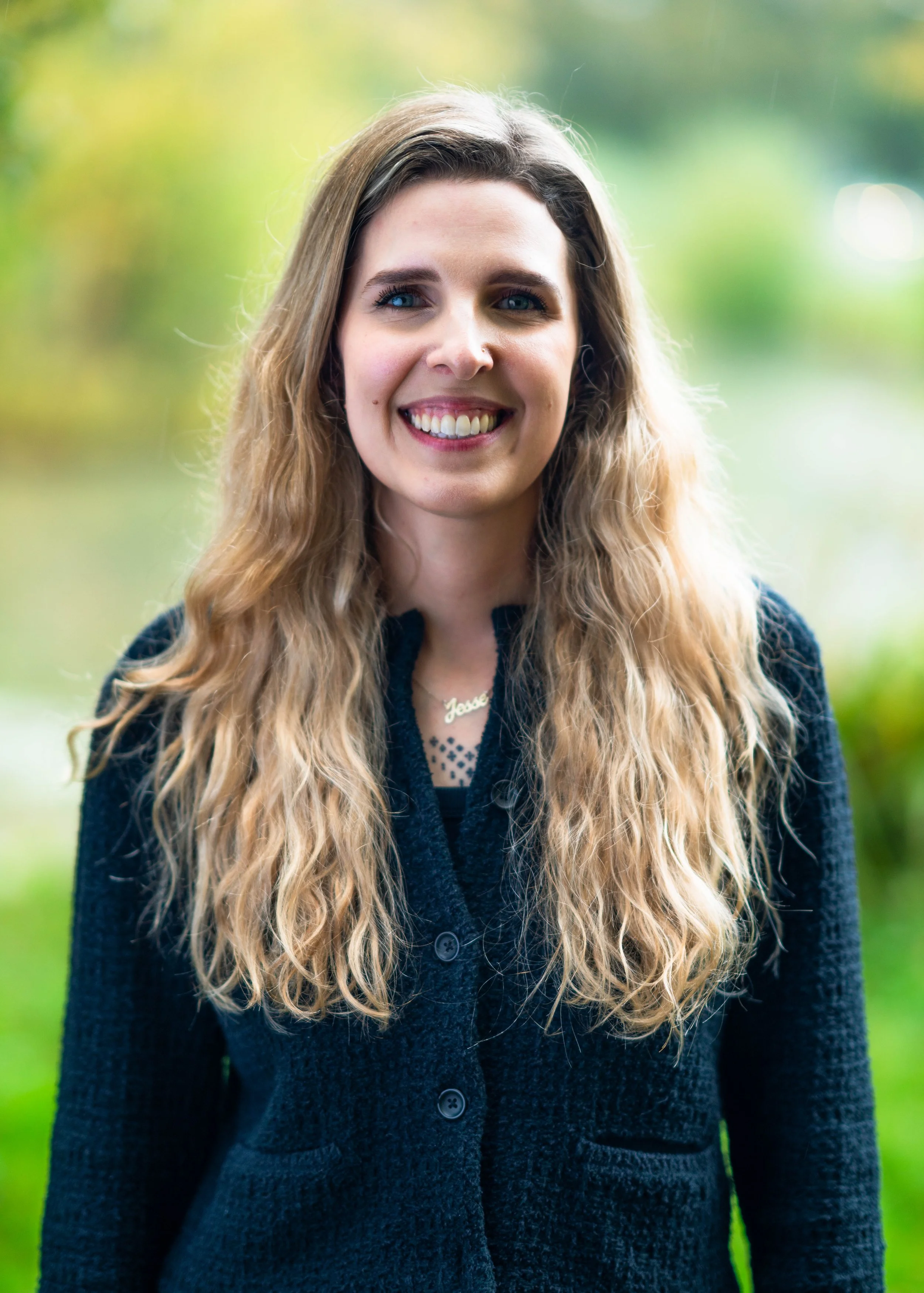 A young woman with long, wavy blonde hair smiling outdoors in front of a blurred background of greenery.
