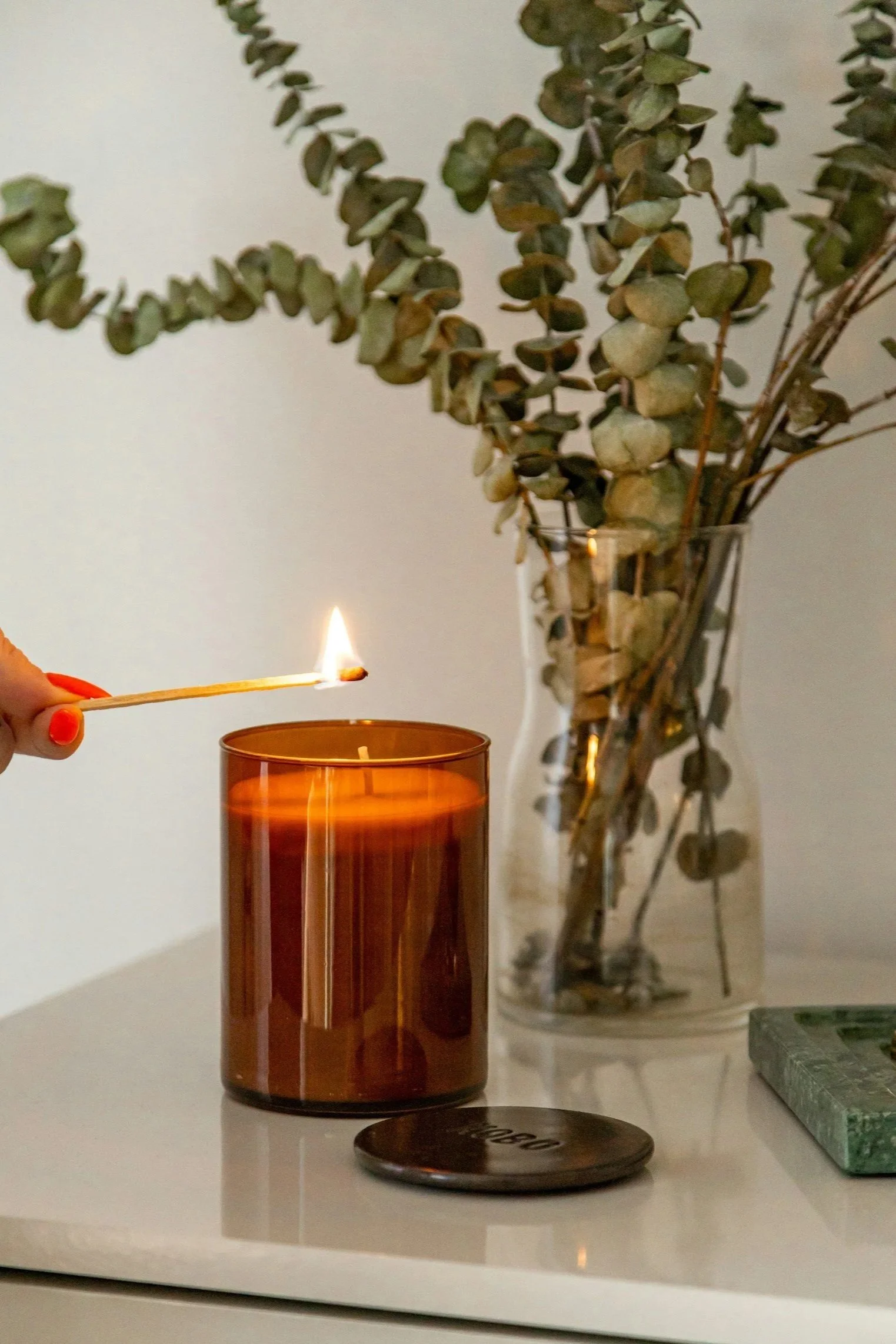 A person is lighting a candle with a matchstick. The candle is in a brown glass container on a white surface. Behind it is a tall glass vase with dried eucalyptus branches. There is also a small round object and a rectangular green stone on the surface.