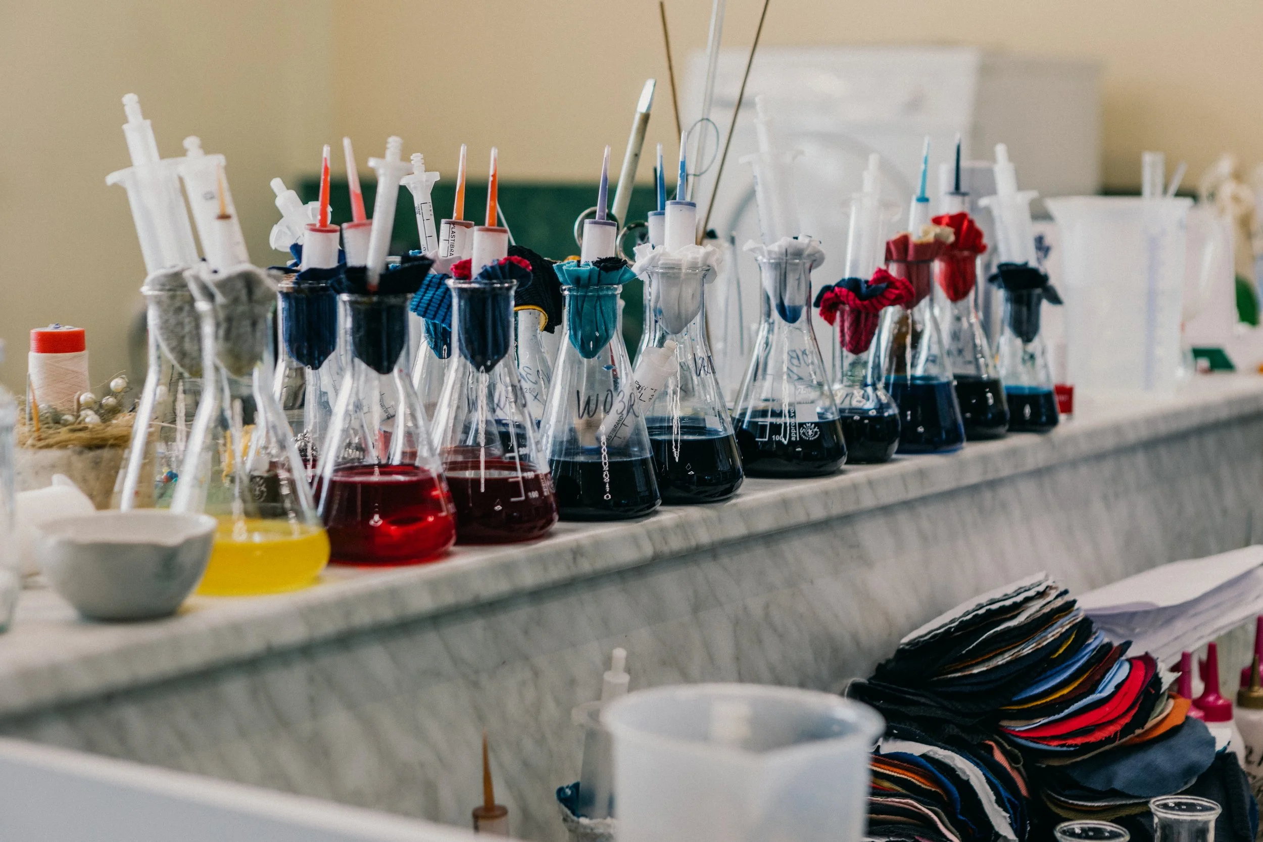 Laboratory setup with multiple glass flasks containing various colored liquids, glass pipettes, and cloth pieces attached to the pipettes, all arranged on a countertop.