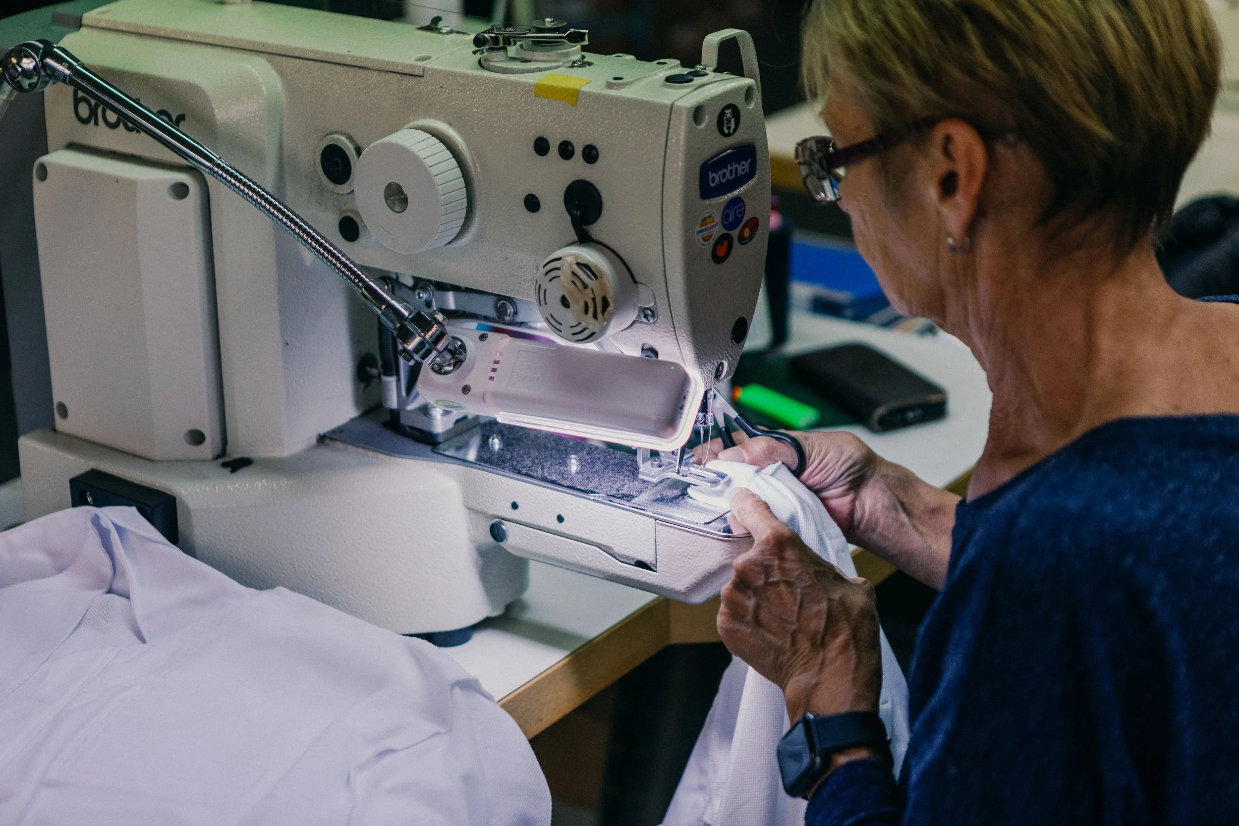 An elderly woman sewing white fabric on a large industrial sewing machine.