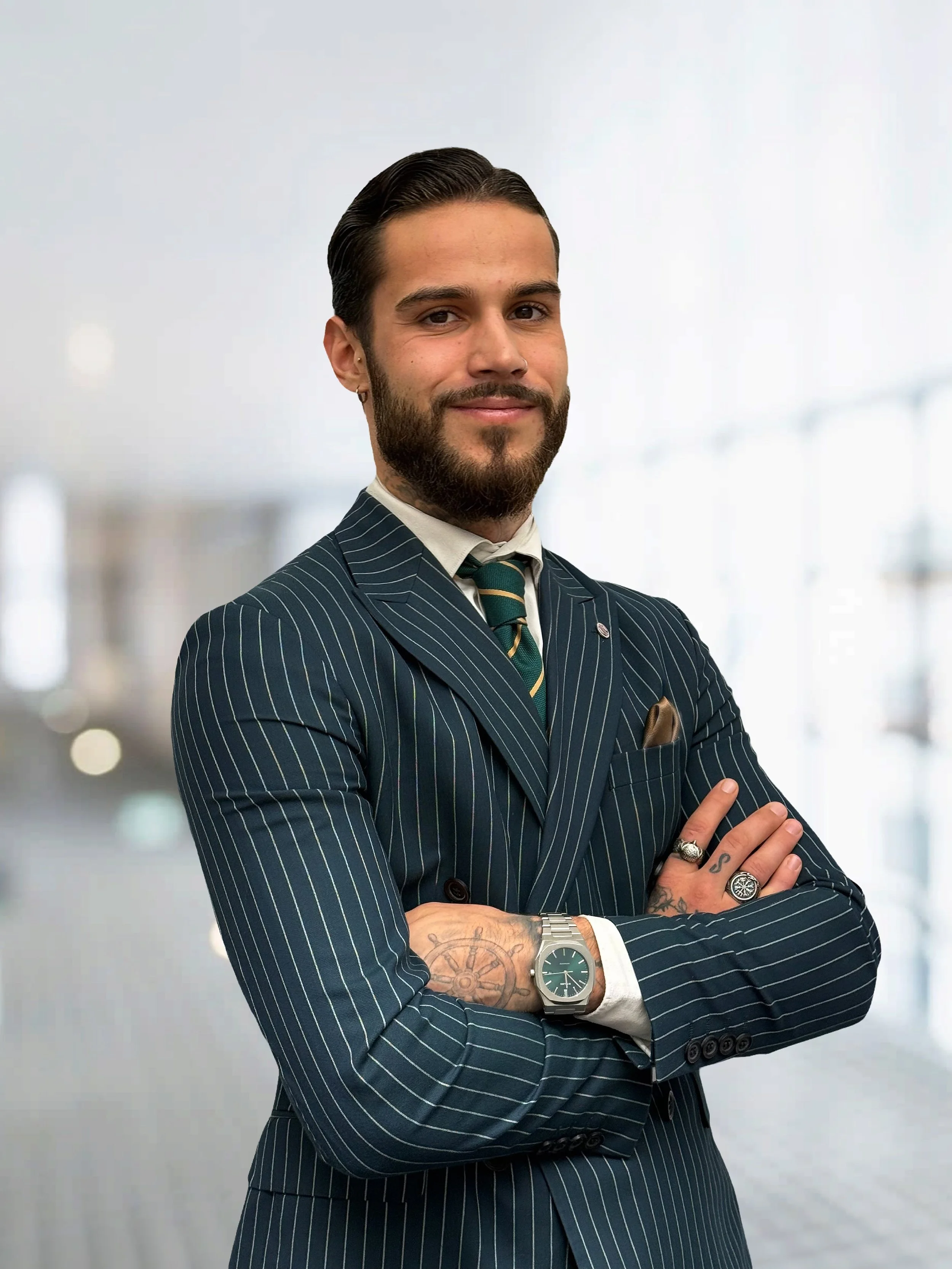 Ein Mann in Business-Kleidung mit dunklem gestreiftem Anzug, Krawatte und Armbanduhr posiert mit verschränkten Armen in einem modernen Büro.