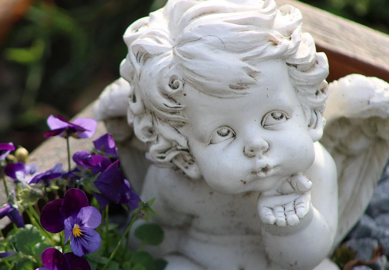 Eine weiße Gartenzierfigur eines kleinen Kindes mit flauschigem Haar und einem nachdenklichen Gesichtsausdruck, umgeben von violetten Blumen.