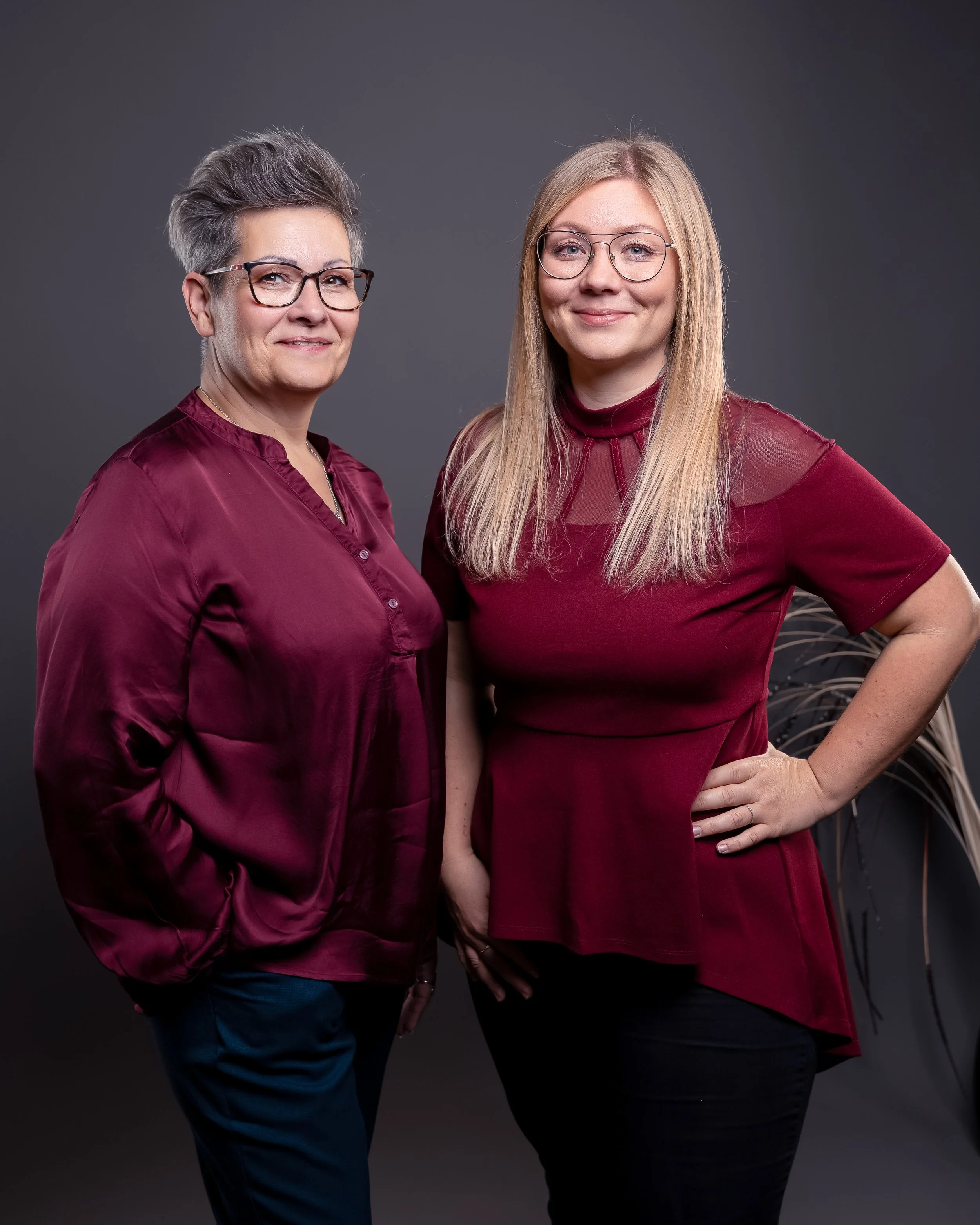 Zwei Frauen in Rot vor dunklem Hintergrund, eine ältere mit grauem kurzen Haar, die andere jüngere mit langem blondem Haar, beide tragen Brillen.