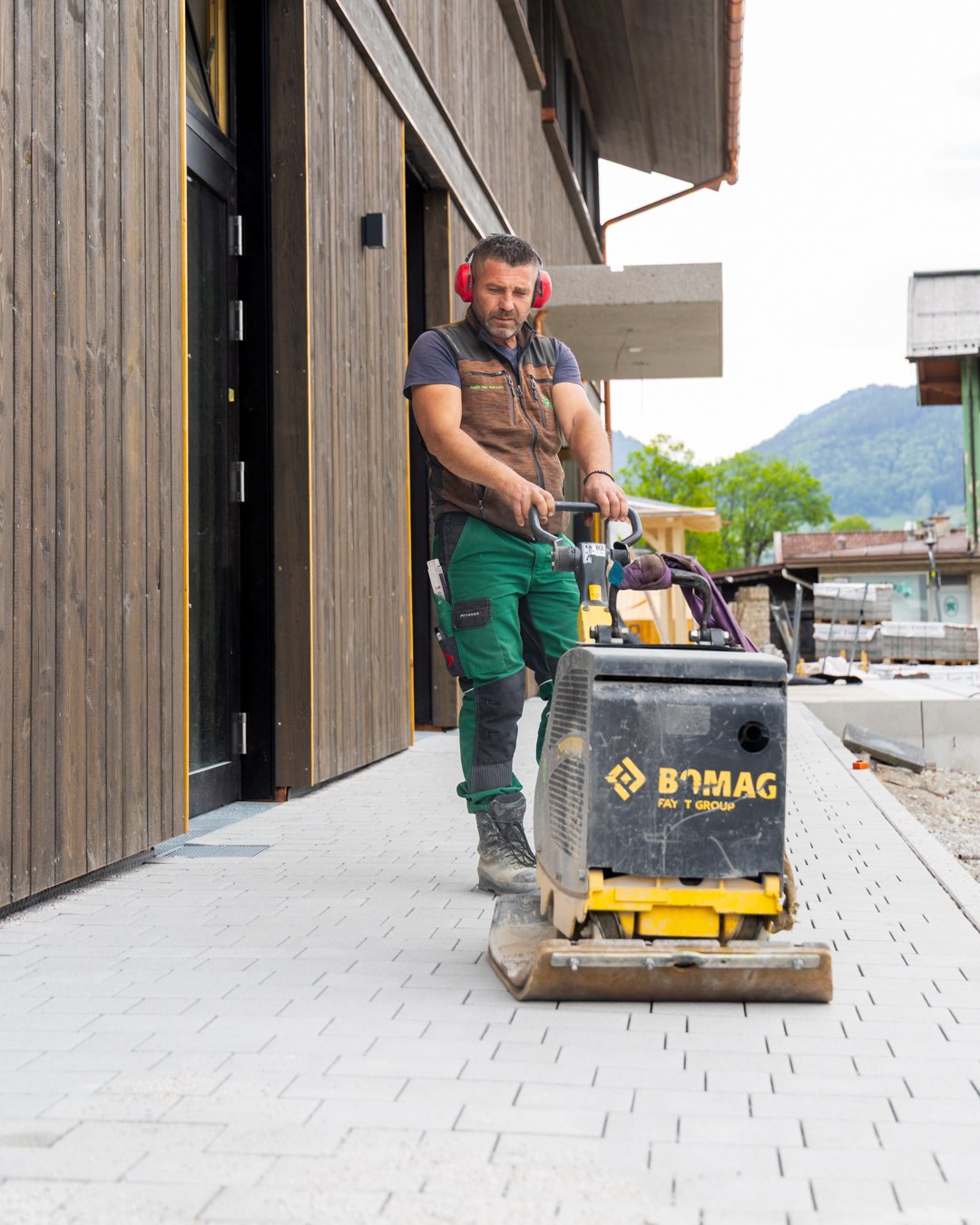 Bauarbeiter bei der Arbeit mit Rüttelplatte auf einer gepflasterten Baustelle vor einem Holzhaus mit bergigem Hintergrund.