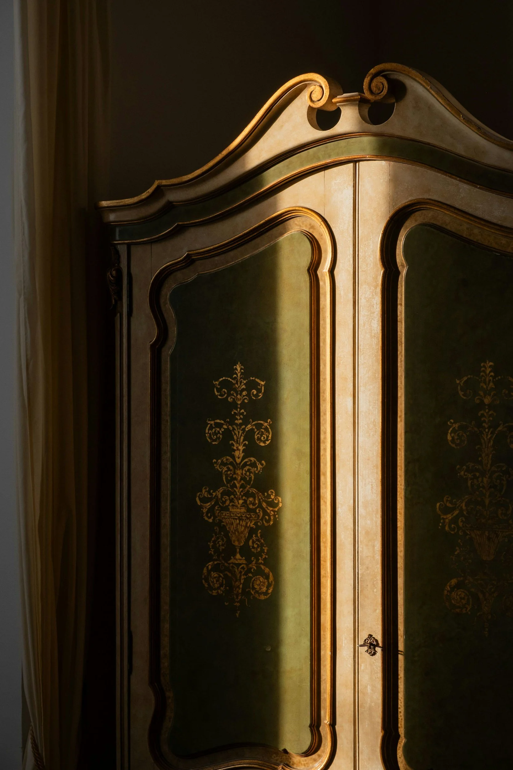 Close-up of an antique wooden wardrobe with ornate gold detailing and green panels, partially illuminated by natural light.
