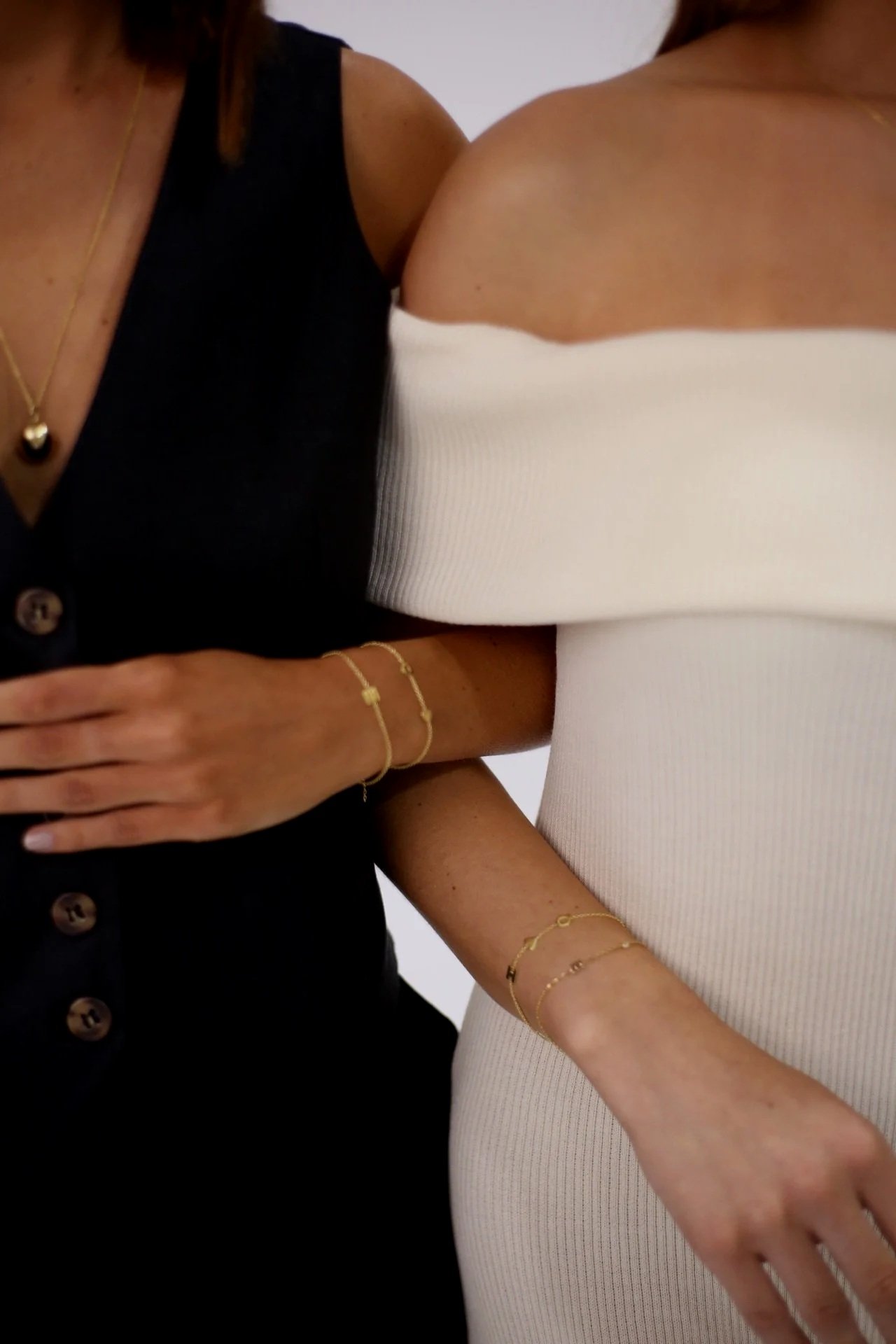 Close-up of two women standing together, one in a black sleeveless top and the other in an off-the-shoulder white dress, both wearing gold jewelry.