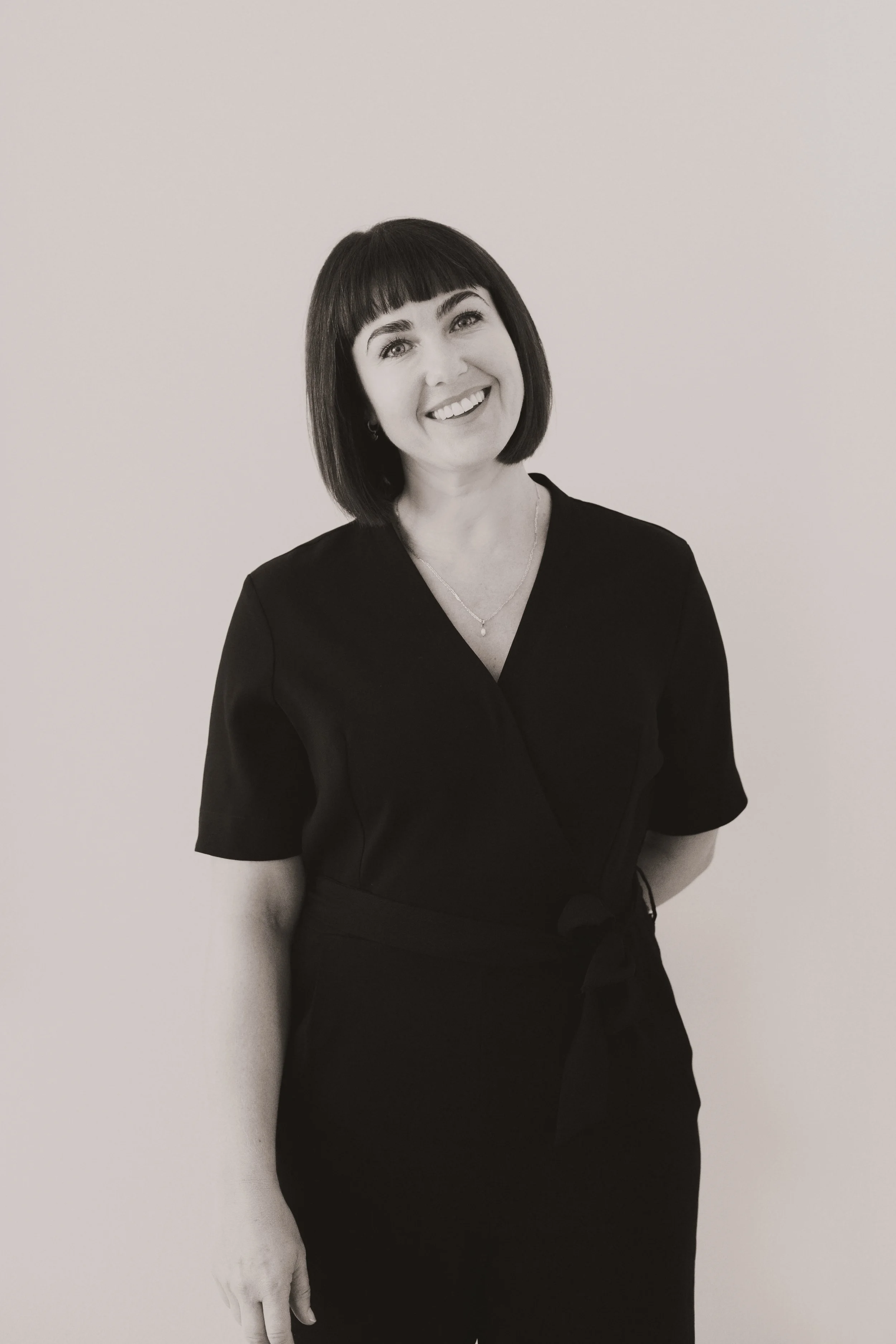 A woman with dark, straight, bob hairstyle and bangs, smiling while wearing a dark V-neck top and a necklace, standing in front of a plain white wall.
