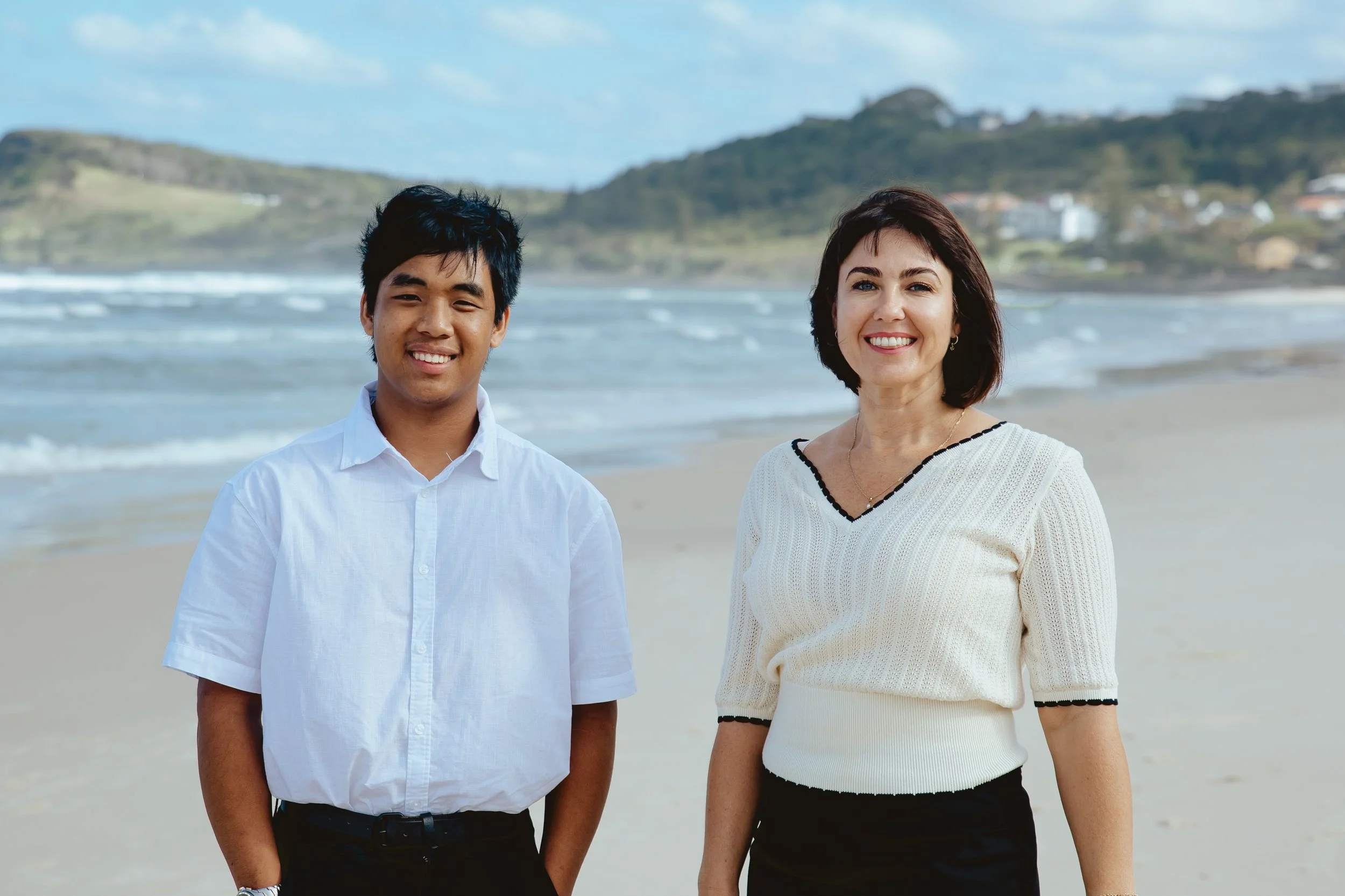 A young man and a woman standing on a beach with ocean waves, hills, and houses in the background, smiling at the camera.