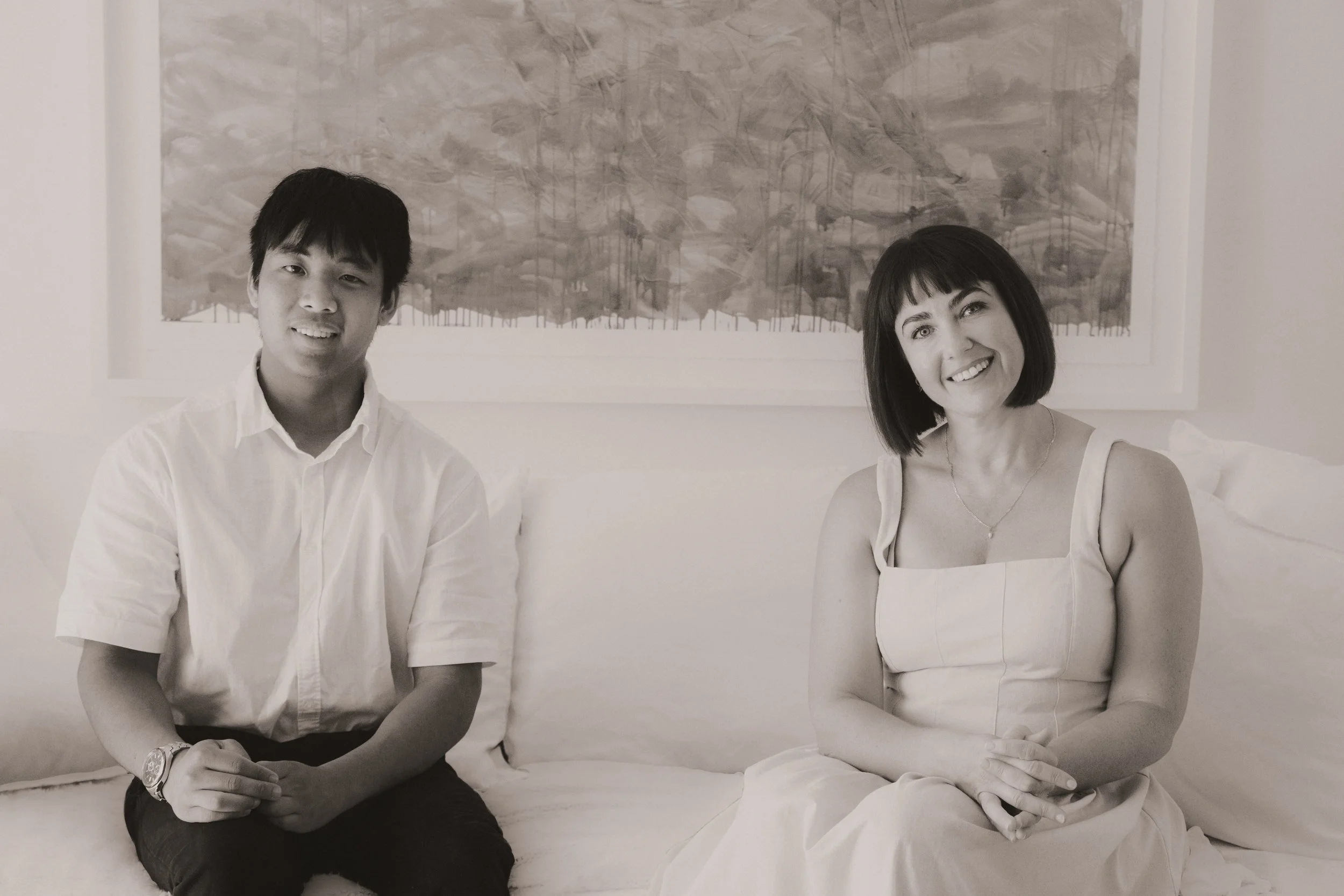 A young man and a woman sitting on a sofa with a framed painting behind them in a well-lit room.