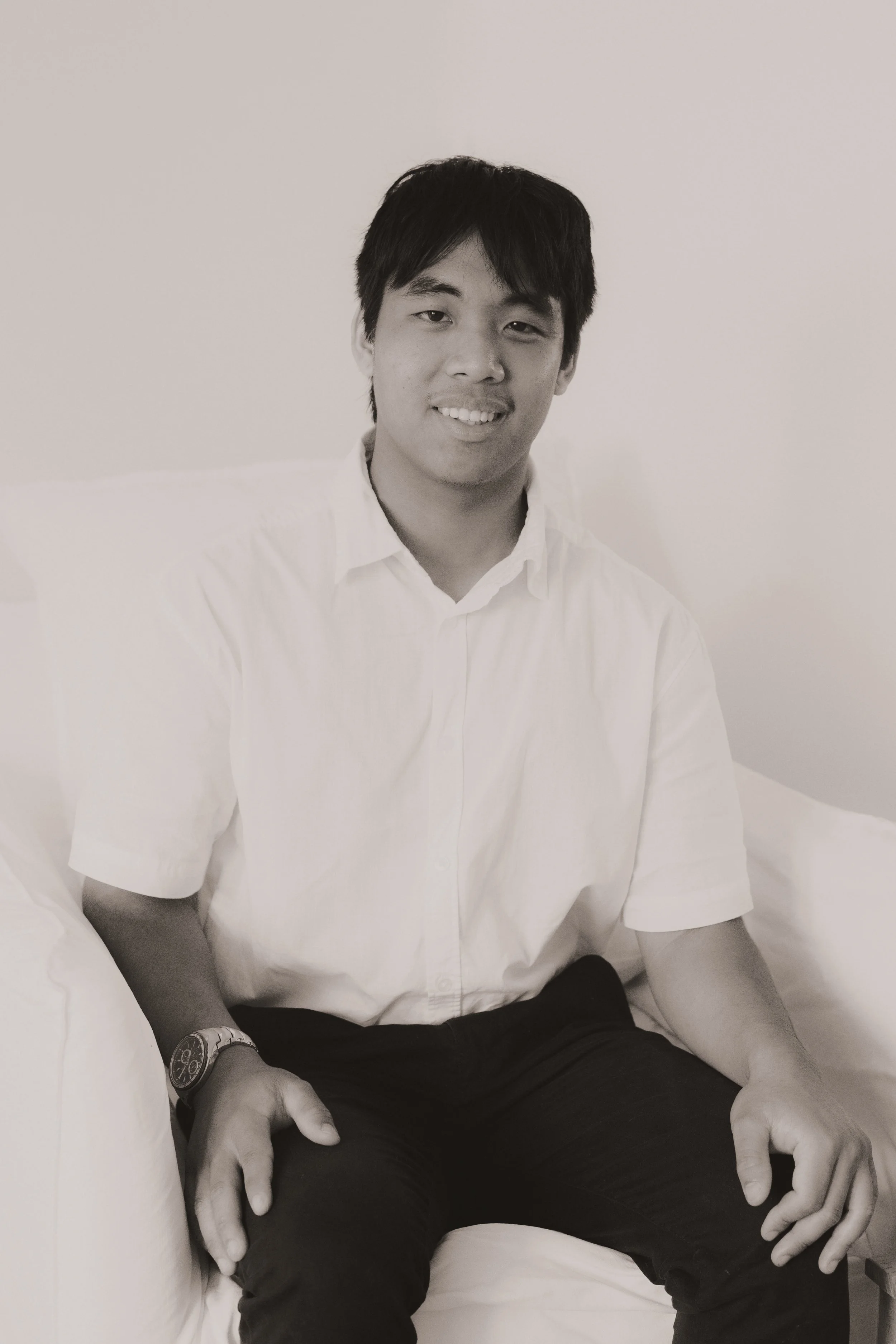 A young man sitting on a bed, smiling, dressed in a white shirt and dark pants, wearing a watch on his left wrist, with a plain wall in the background.