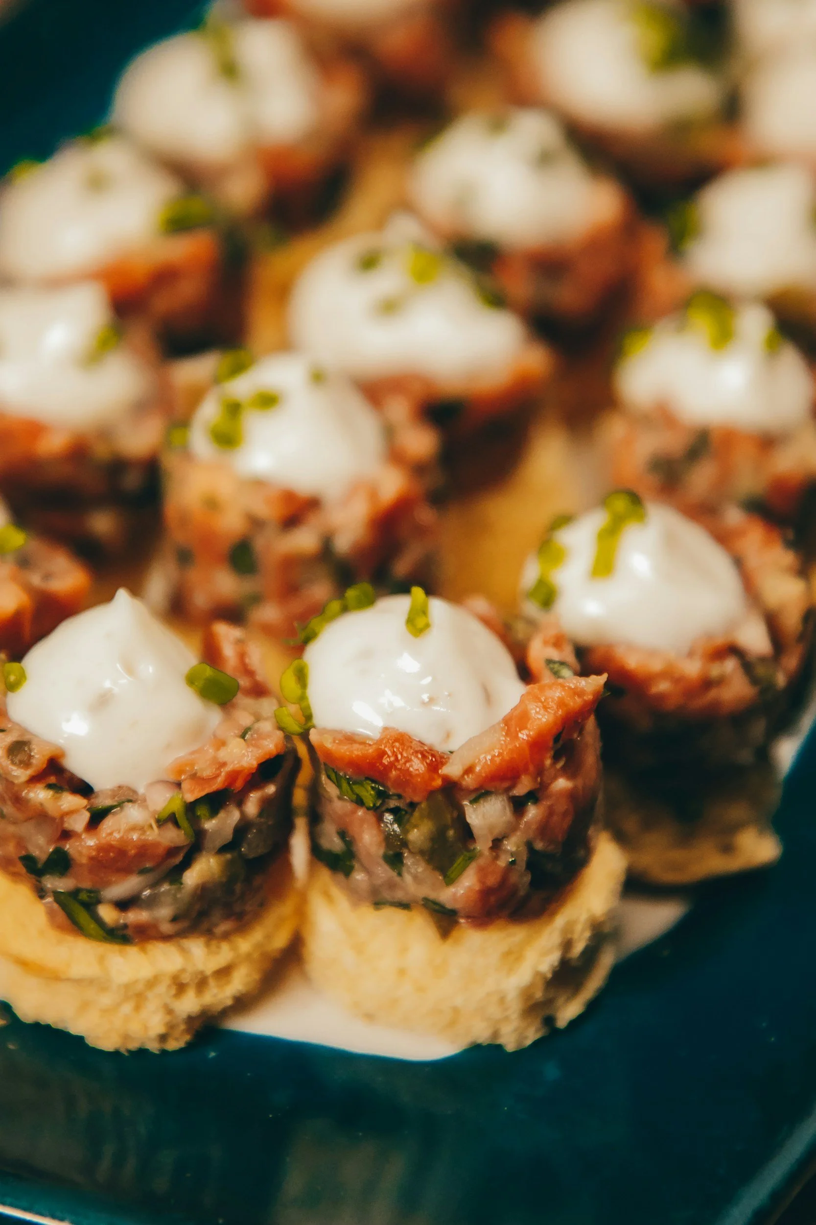 Close-up of savory appetizers topped with creamy white sauce and chopped herbs on a dark plate.