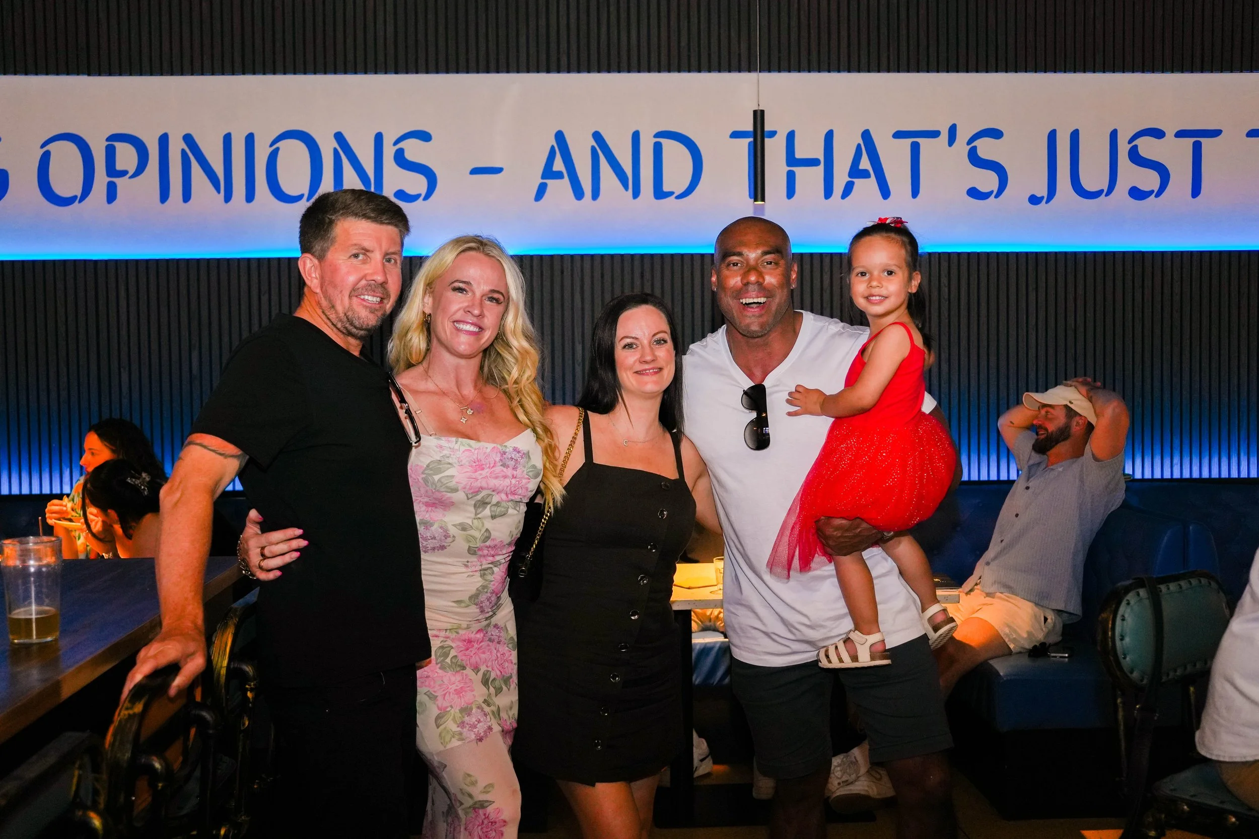 A group of five people posing together at a social gathering. There are three women and two men, with one man holding a young girl dressed in red. In the background, there are two people sitting at a table and a man sitting with his back against the 