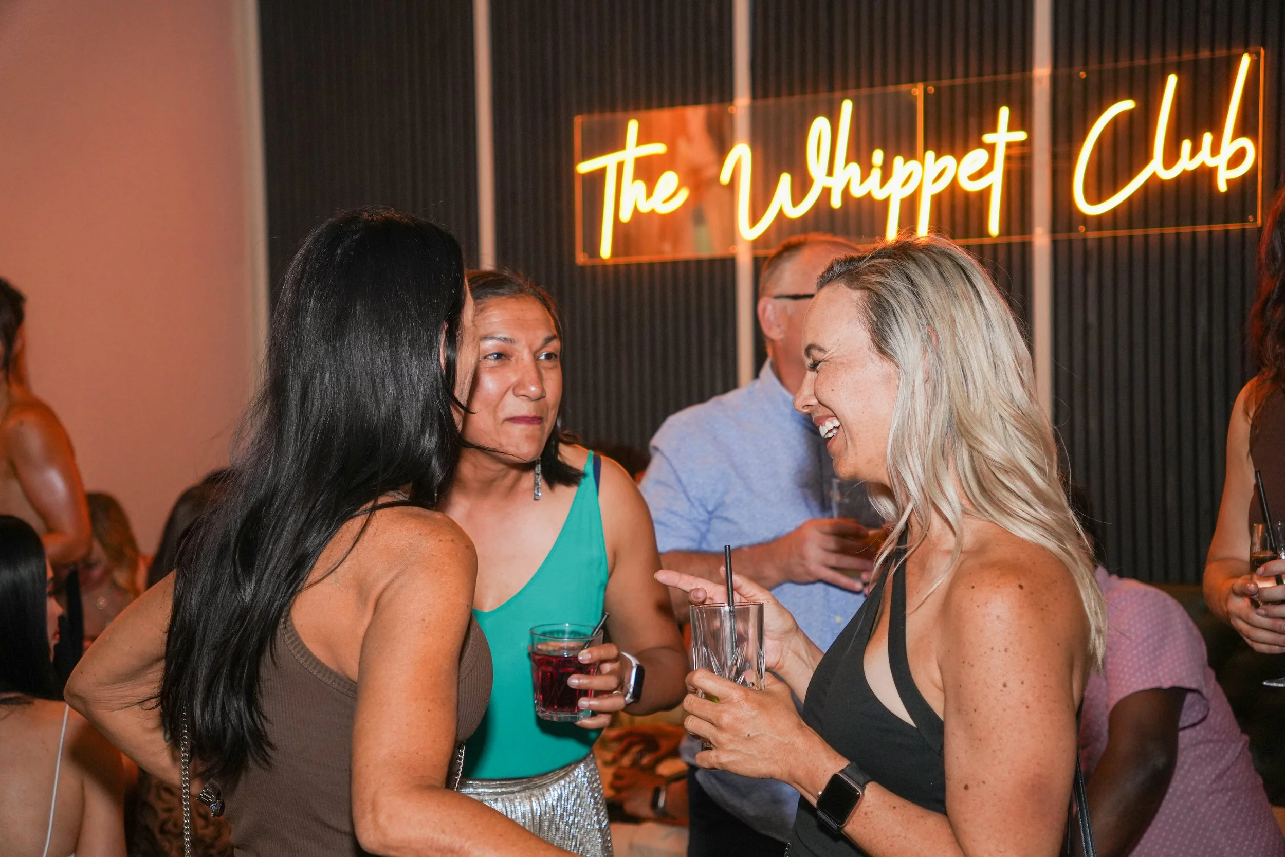 Group of people socializing at a bar with a neon sign that reads 'The Whippet Club' in the background