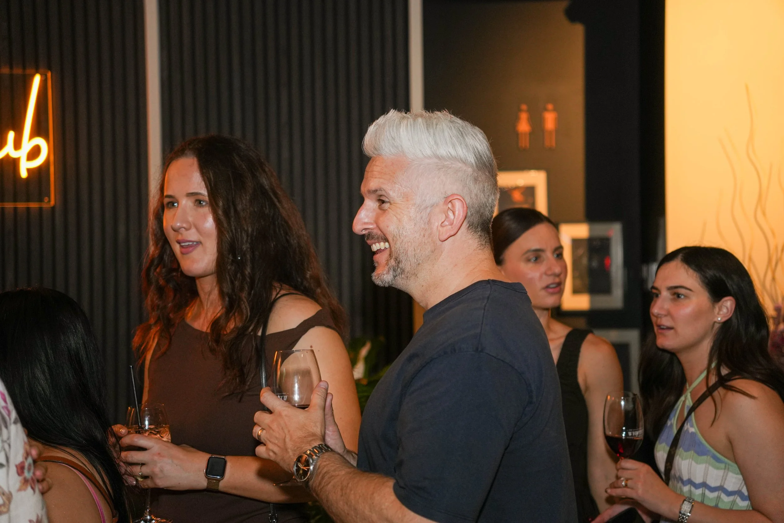 People socializing at a bar or lounge, holding glasses of wine and engaging in conversation. The setting appears casual and lively, with dark walls and warm lighting.