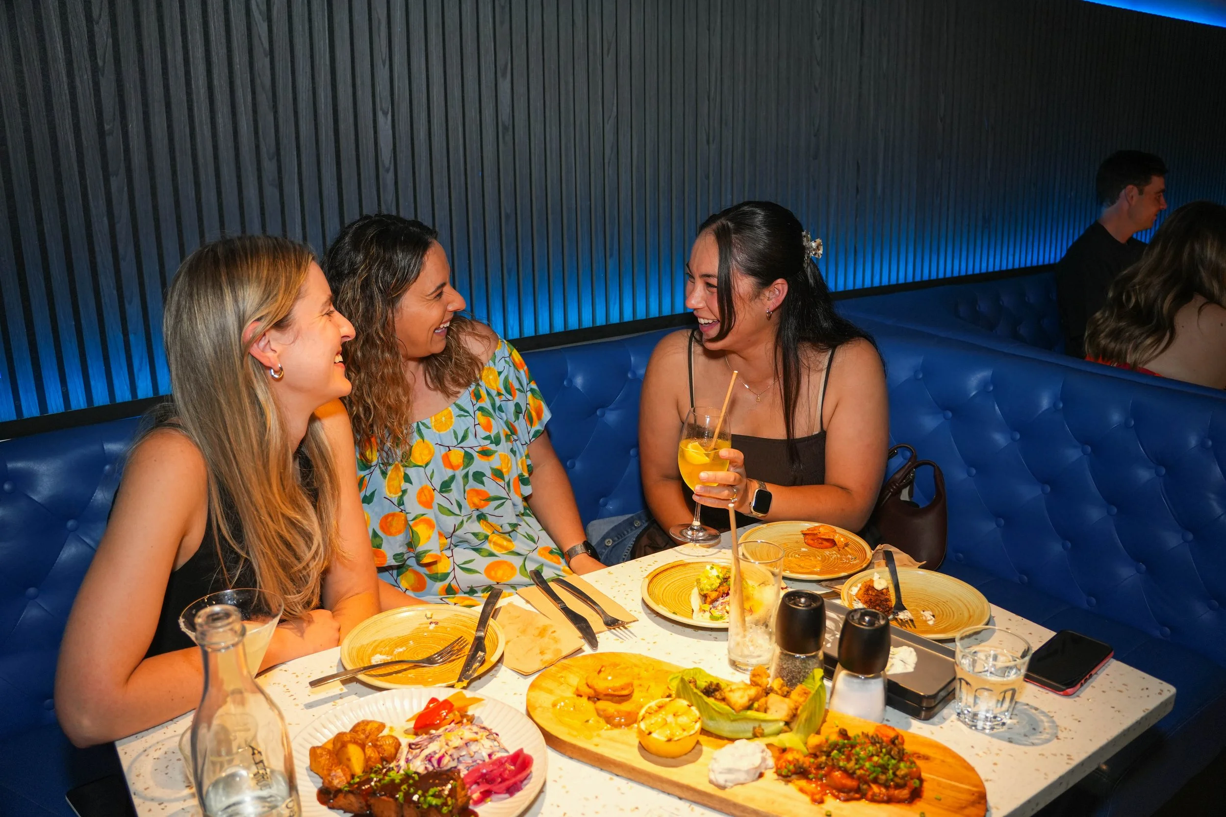 Four women sitting at a table in a restaurant, laughing and talking, with party food and drinks on the table.