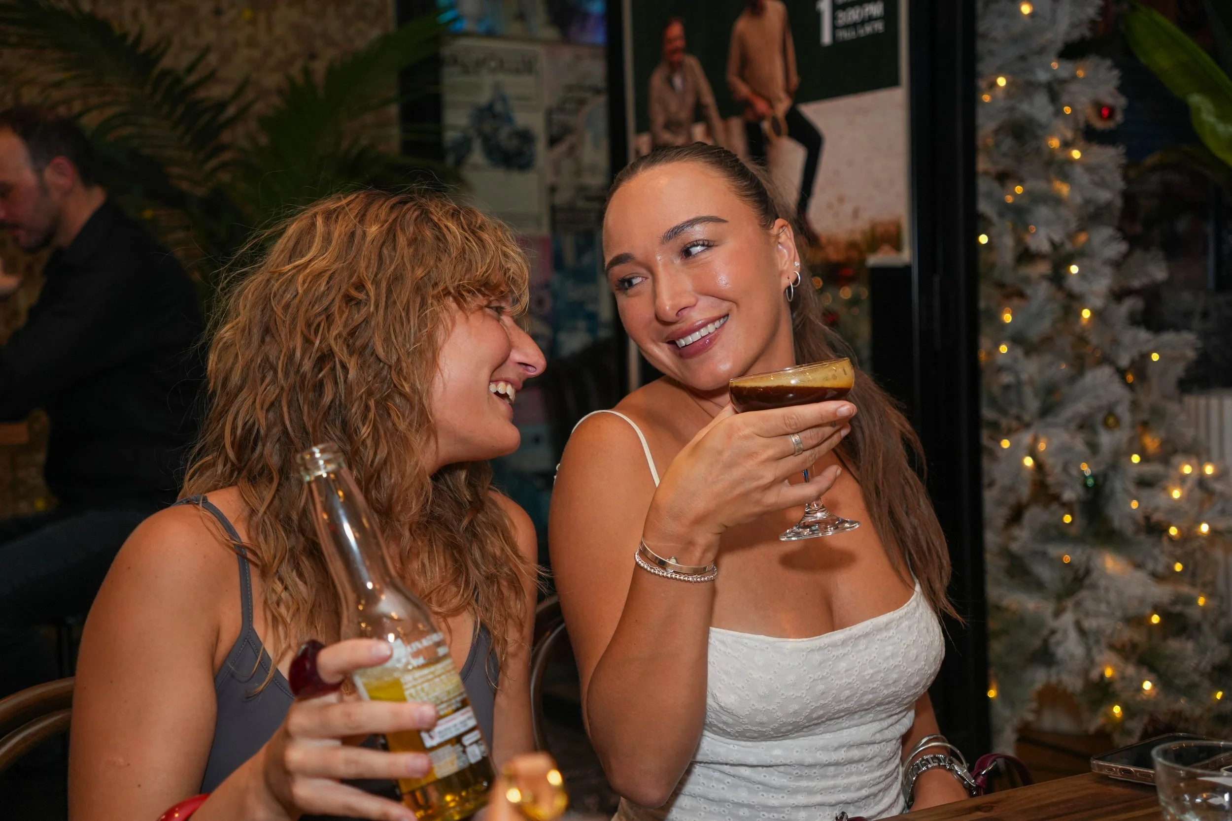 Two women are smiling and enjoying drinks at a social gathering, with festive decorations like a Christmas tree in the background.