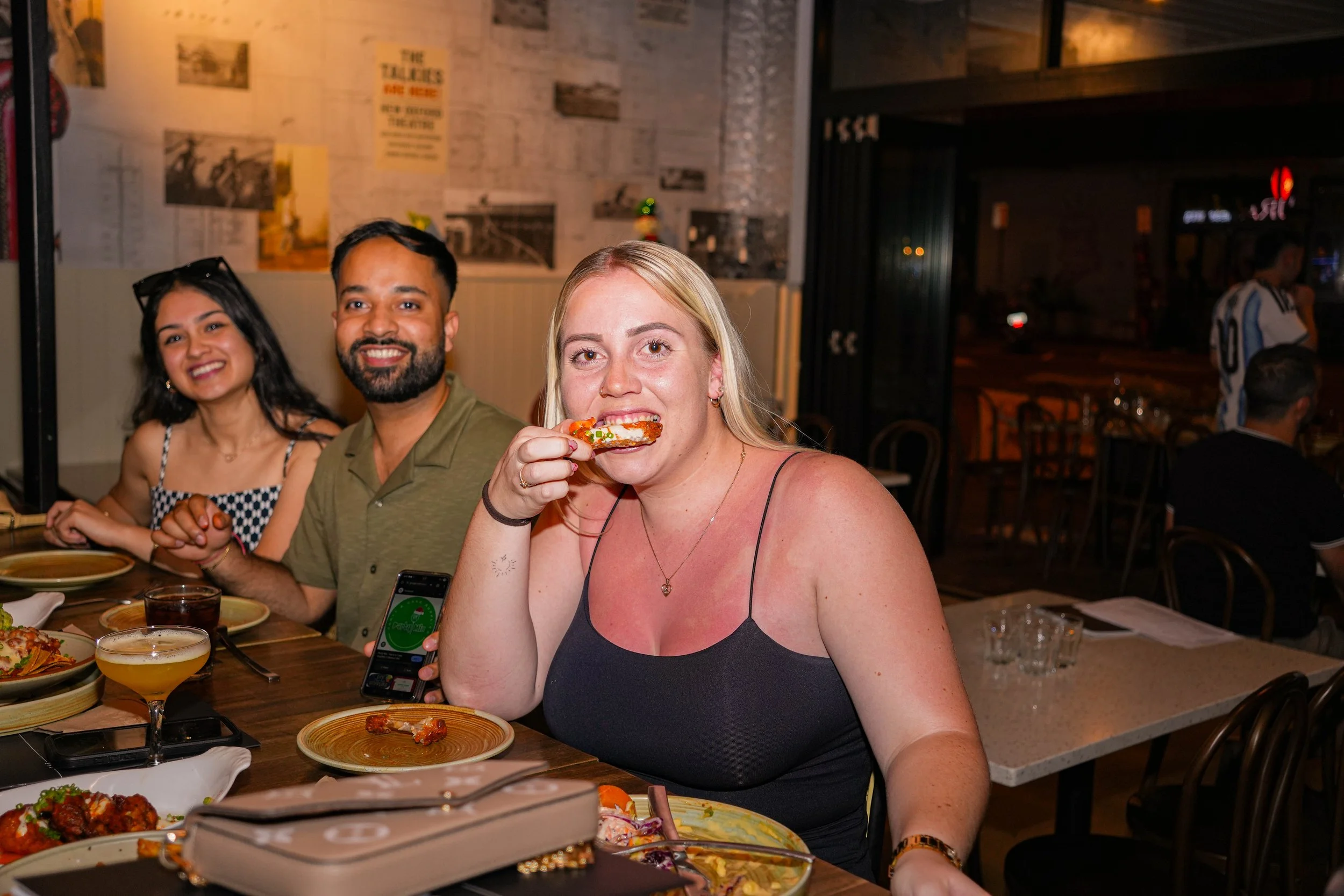 Group of three people dining at a restaurant, with a woman in the foreground taking a bite of food, others smiling at the camera, with plates of food and drinks on the table.