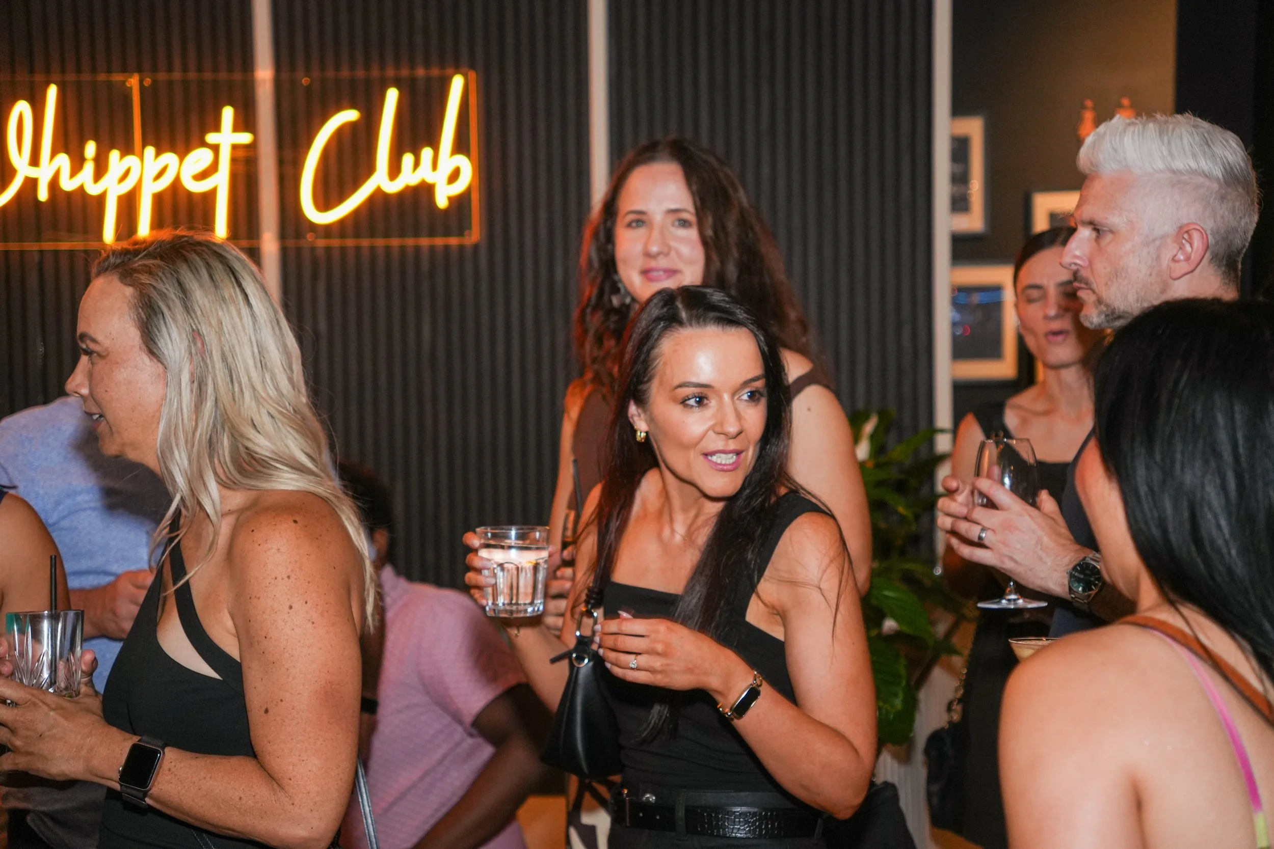Group of people socializing at an art club event, with a neon sign that reads 'Shippet Club' in the background.