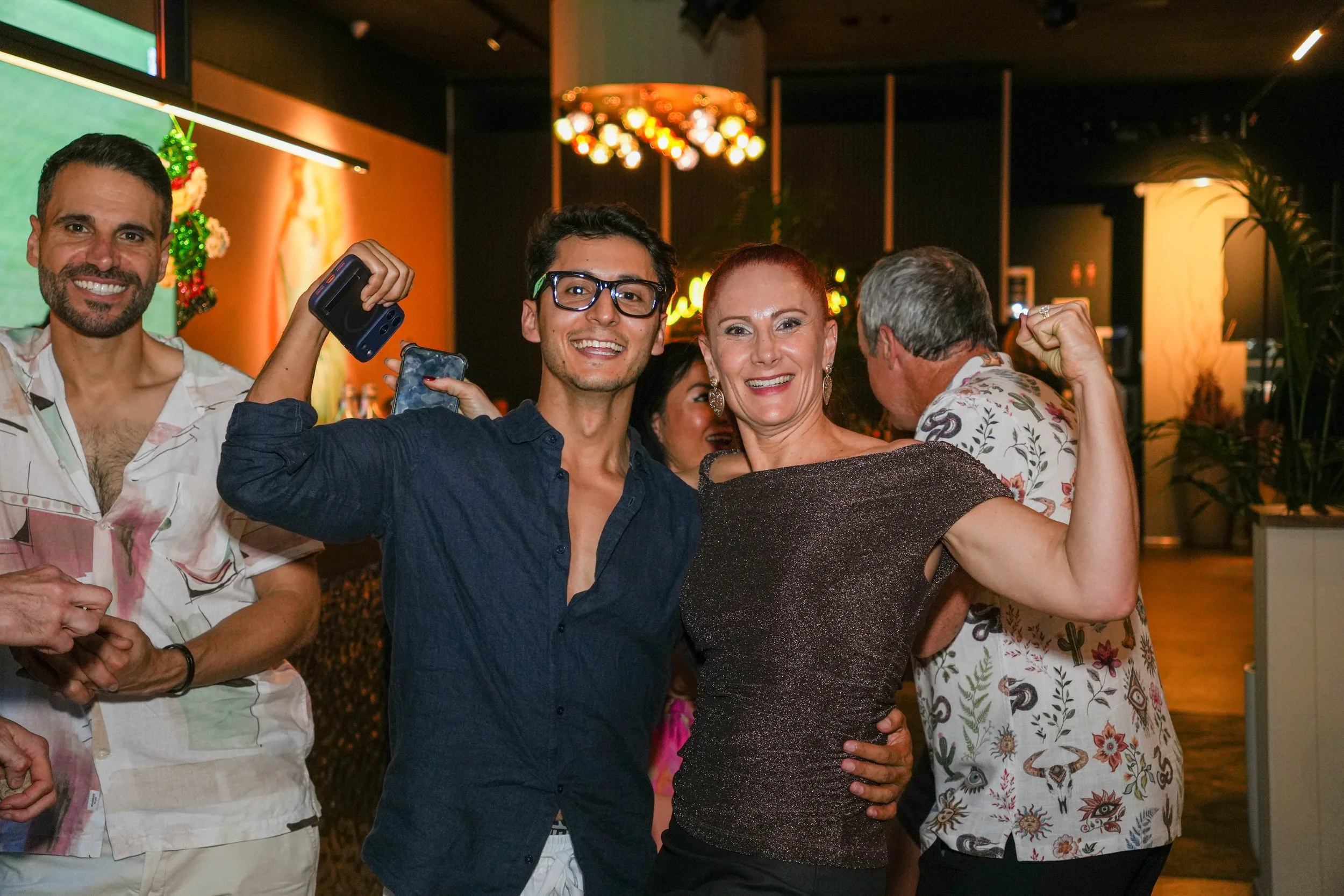 Group of people at a celebration, with two women in the foreground showing off their muscles and smiling at the camera, in a lively Indoor setting with warm lighting and decorative elements.