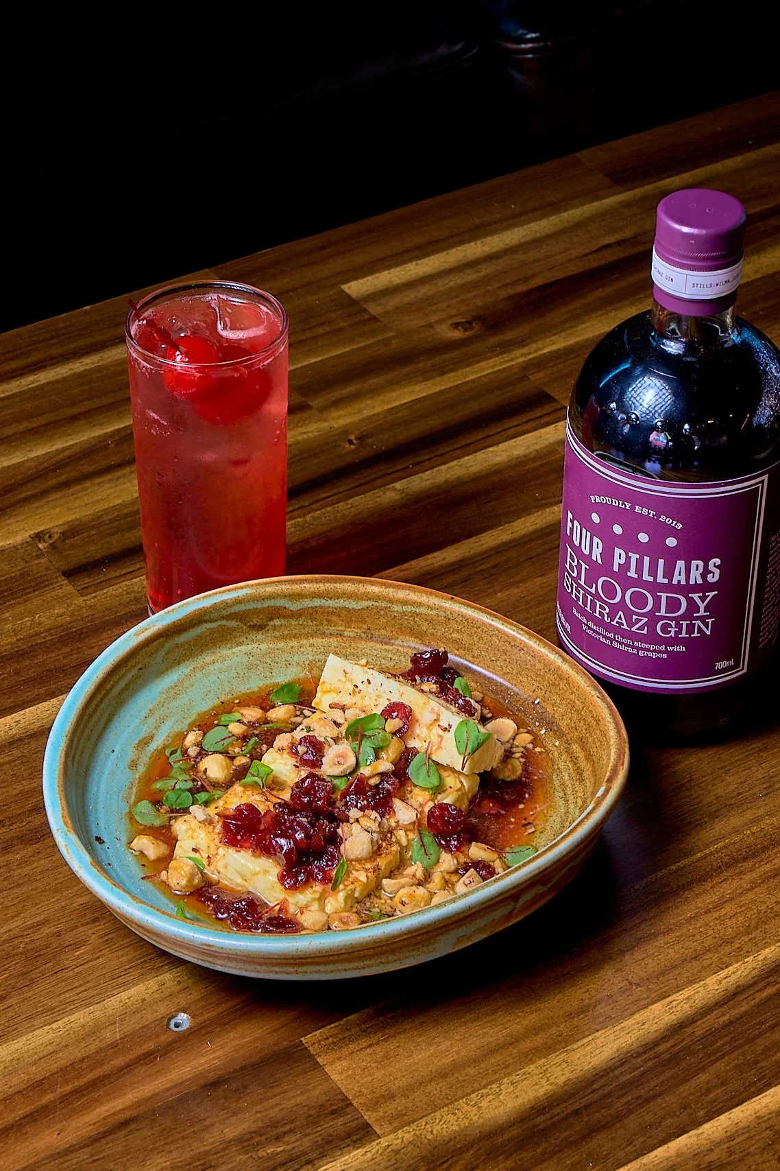A bowl of creamy cheese topped with chopped nuts, dried cranberries, and microgreens, accompanied by a tall glass of pink cocktail with ice and cherries, and a bottle of Four Pillars Bloody Shiraz Gin on a wooden table.