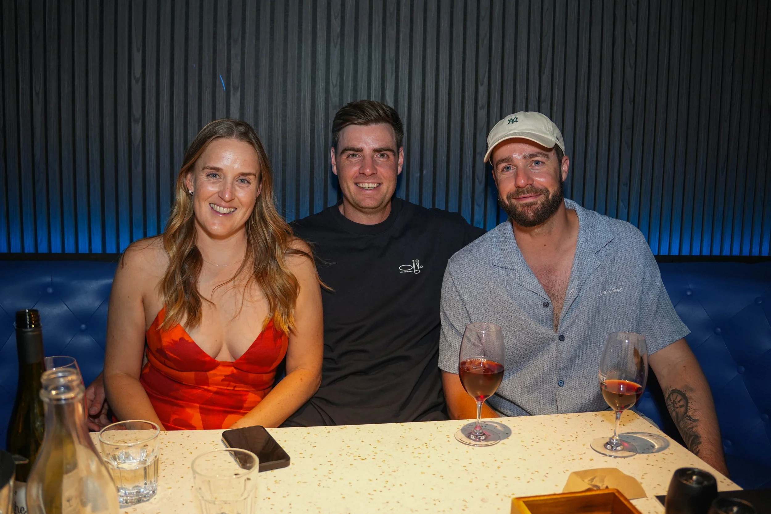 Three people sitting at a dining table, smiling, with wine glasses and drinks on the table, in a modern restaurant setting with dark wall paneling and blue lighting.