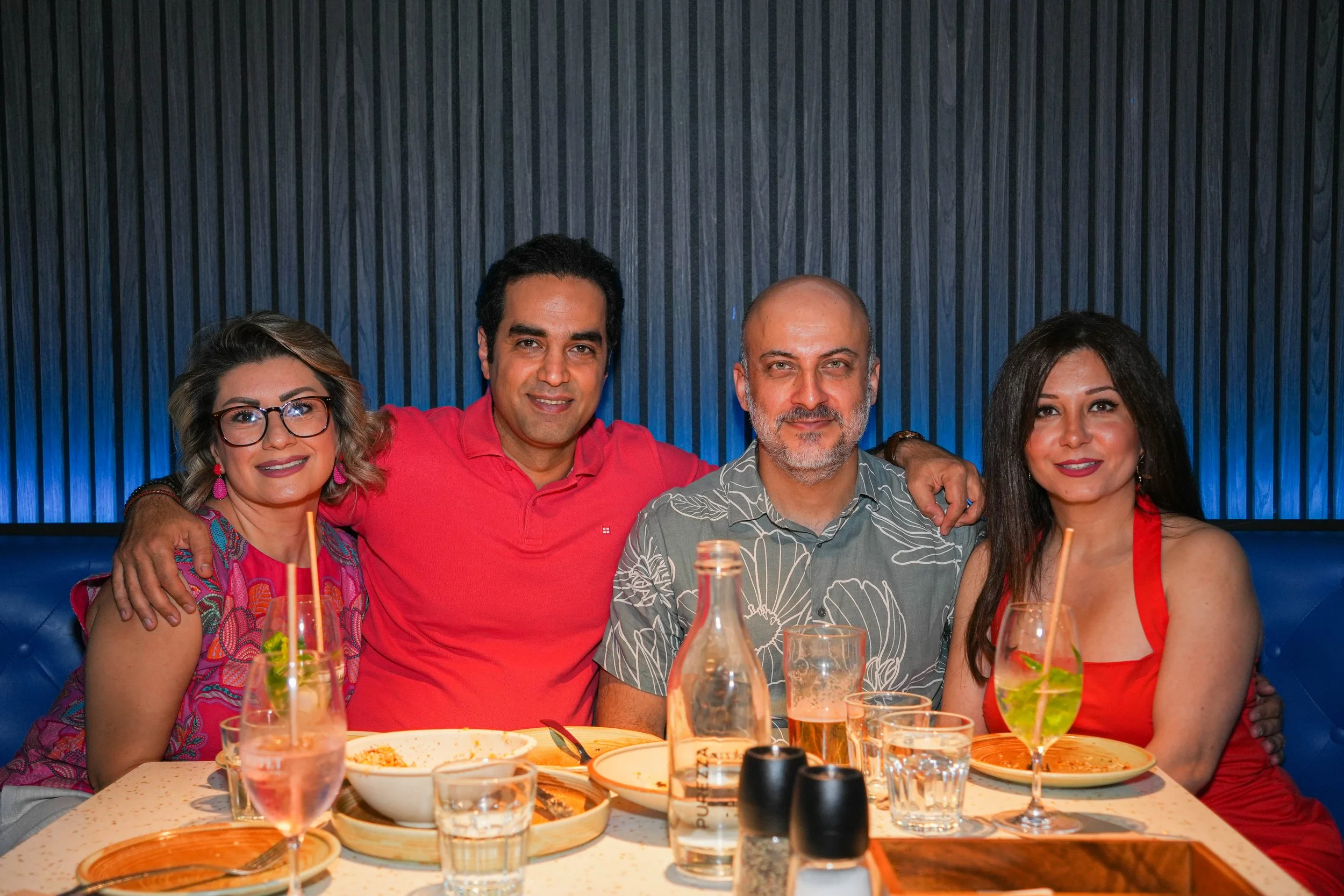 Four people sitting at a dining table during a meal, with plates, drinks, and condiments visible, in a restaurant with a dark striped wall background.