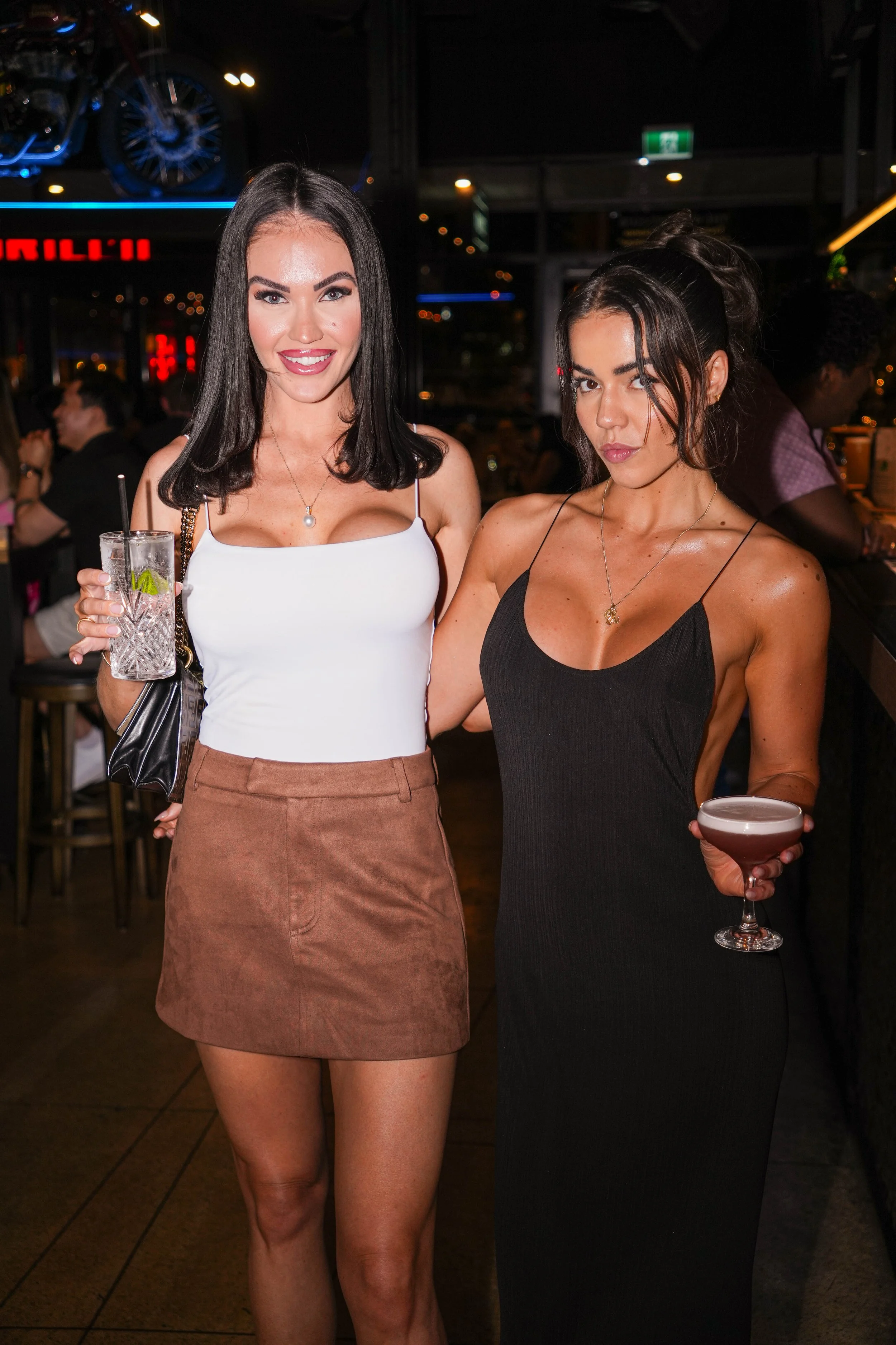 Two women posing at a bar or nightclub, holding drinks, with a dark background and neon lights.