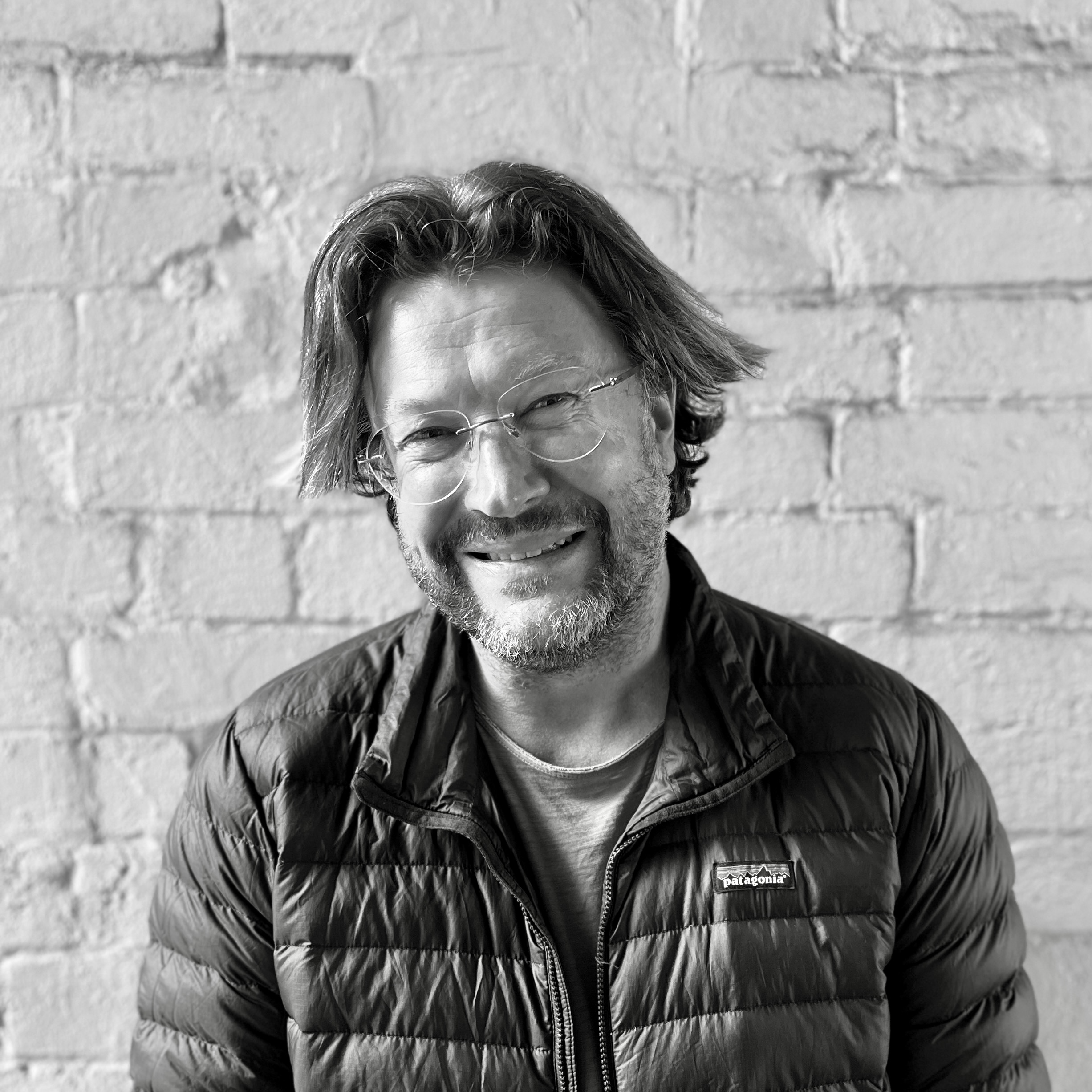 A smiling man with glasses, a beard, and medium-length hair, wearing a Patagonia puffer jacket, sitting in front of a brick wall.