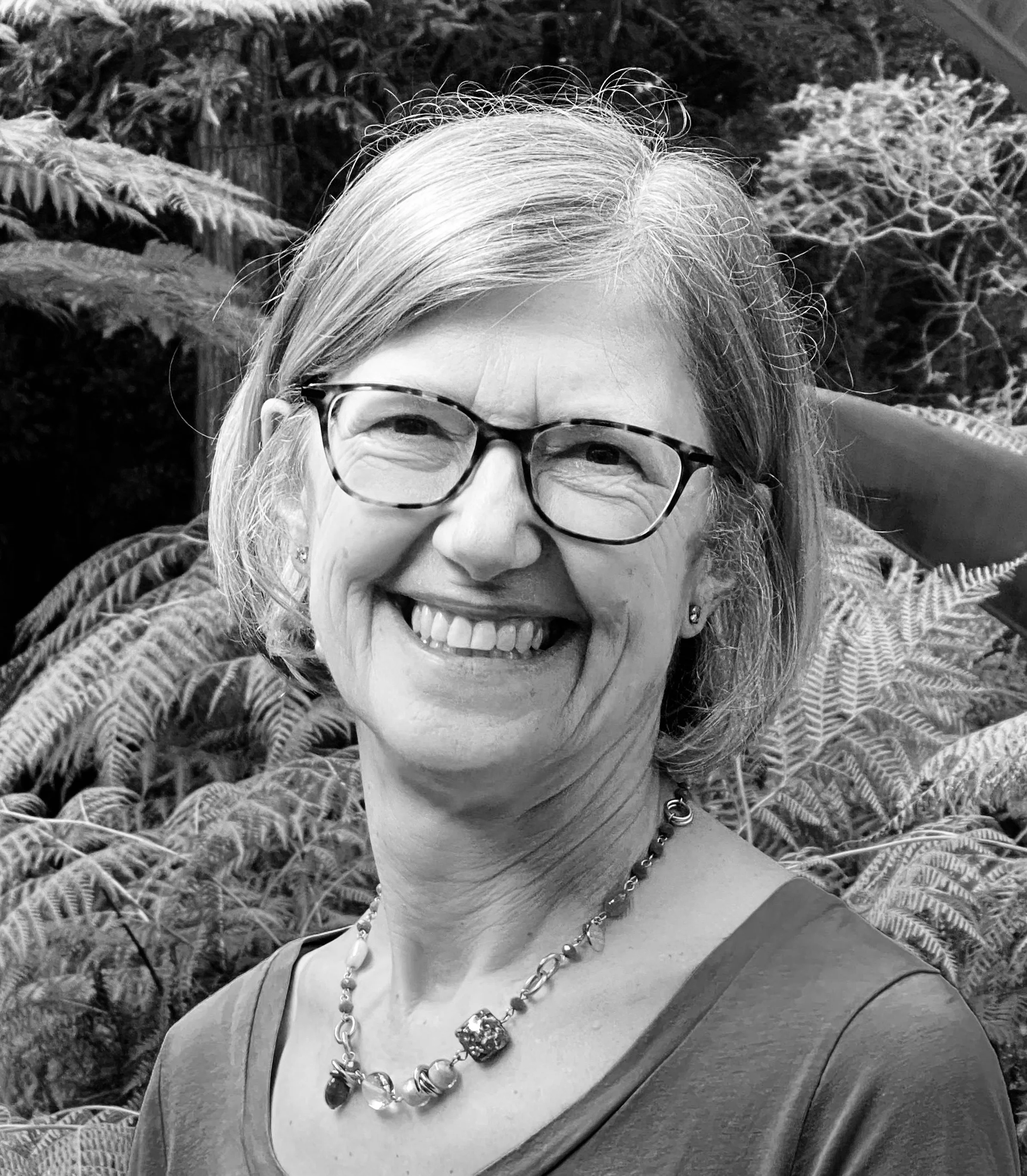 A smiling middle-aged woman with glasses and earrings, wearing a necklace, outdoors with lush foliage in the background.