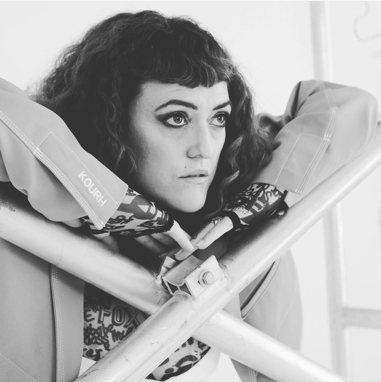 A woman with curly hair resting her chin on her arms, leaning on a metal railing, with a thoughtful expression.
