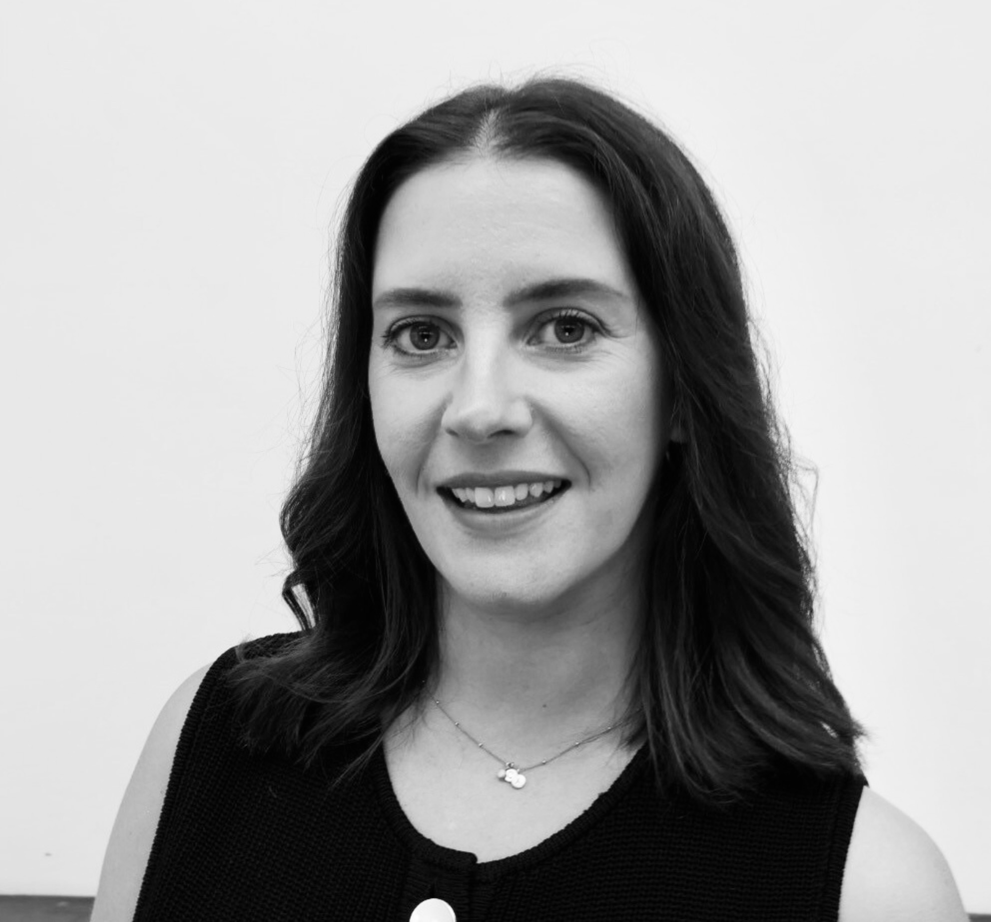 Black-and-white portrait of a woman with long wavy hair, wearing a sleeveless top and a necklace, smiling against a plain background.