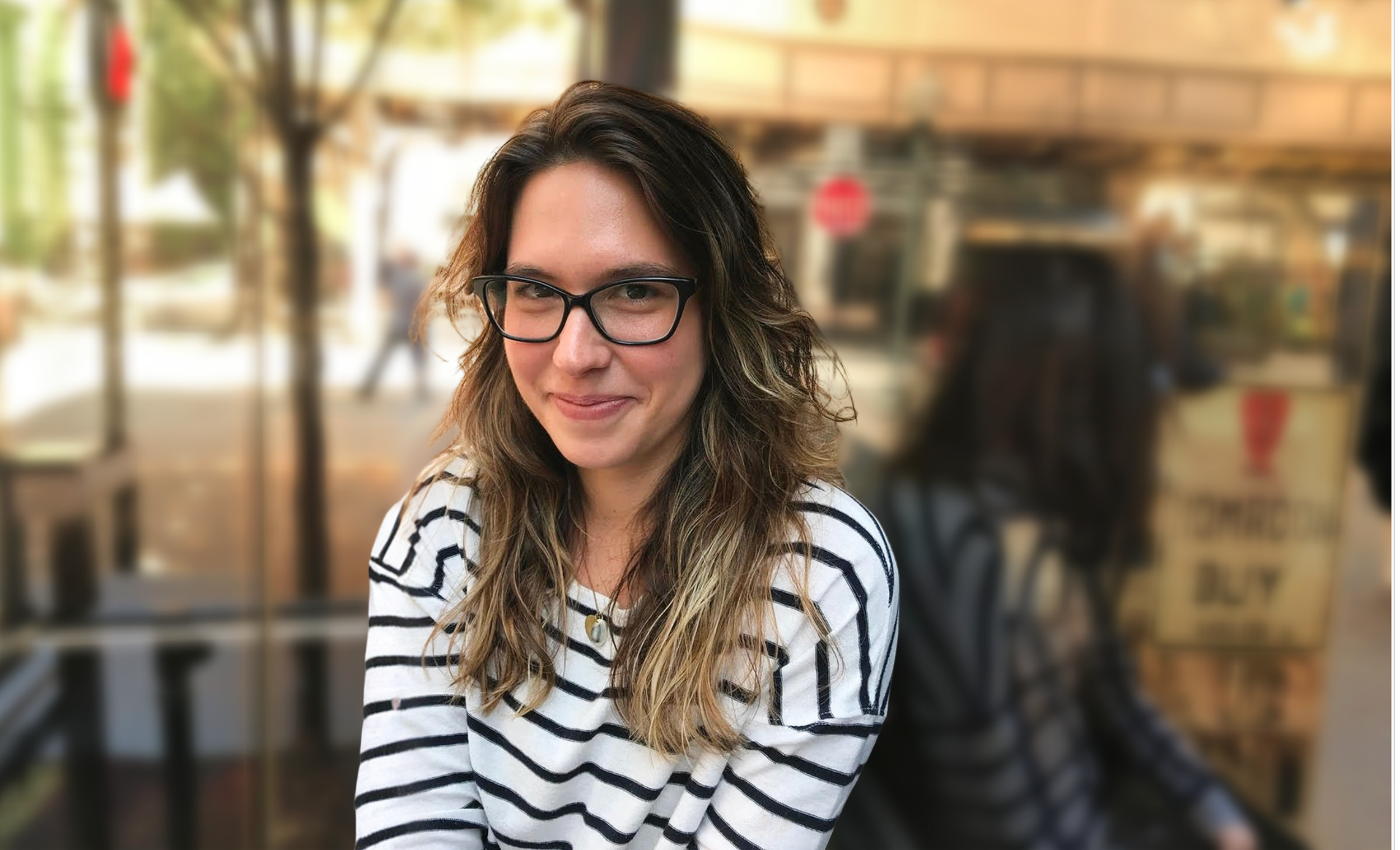 A woman with long wavy hair wearing glasses and a white and black striped shirt, smiling at the camera, sitting outdoors with a blurred background that includes a person and a sign.