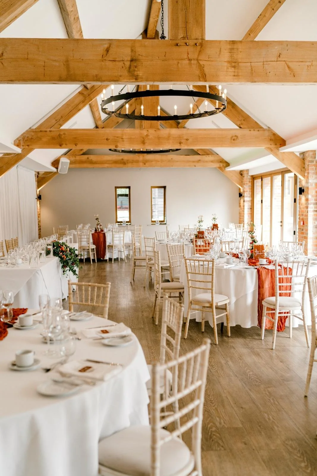 Elegant wedding reception with round tables covered in white tablecloths, decorated with floral centerpieces and orange accents, in a rustic barn converted venue with wood beams, chandelier, and large windows.