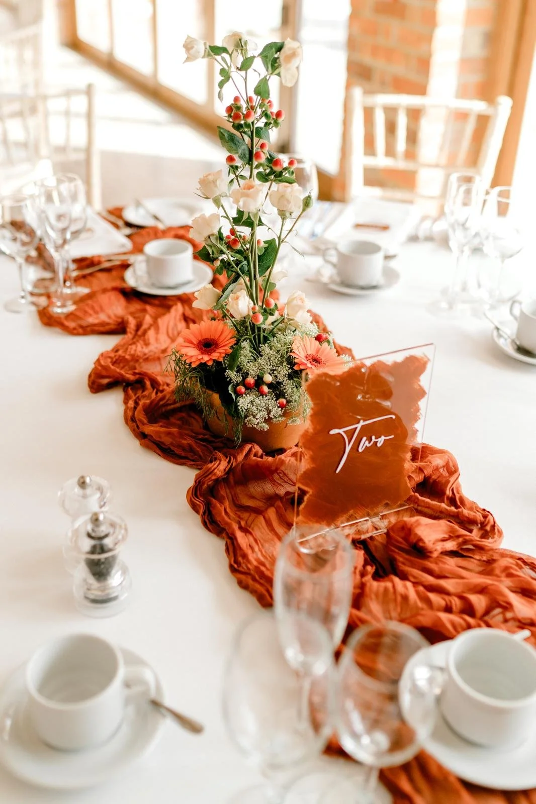 Table decorated with a floral centerpiece featuring white and pink flowers, pink gerbera daisies, and greenery, on a rust-colored fabric runner. Place settings include white cups and saucers, and glasses, with a small glass table number stand labeled 'Two'.