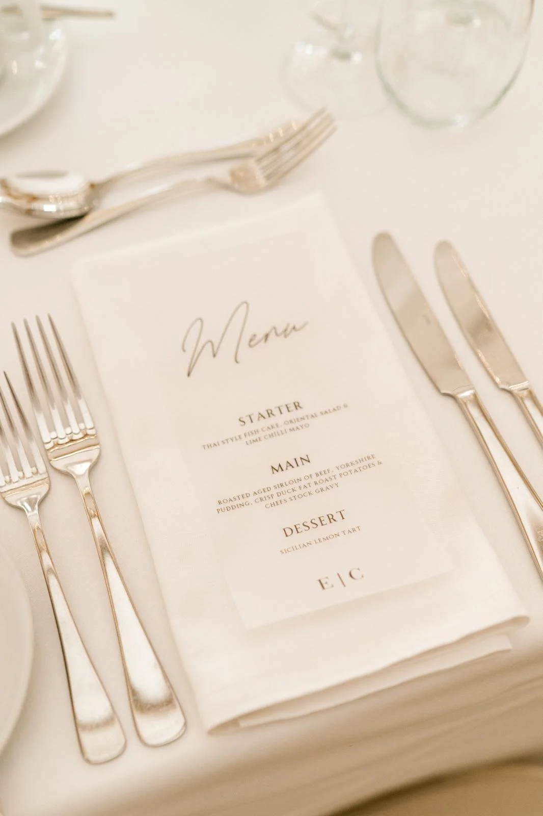 A table setting with a menu on a folded napkin, flanked by silverware including forks, knives, and spoons, with water glasses in the background.
