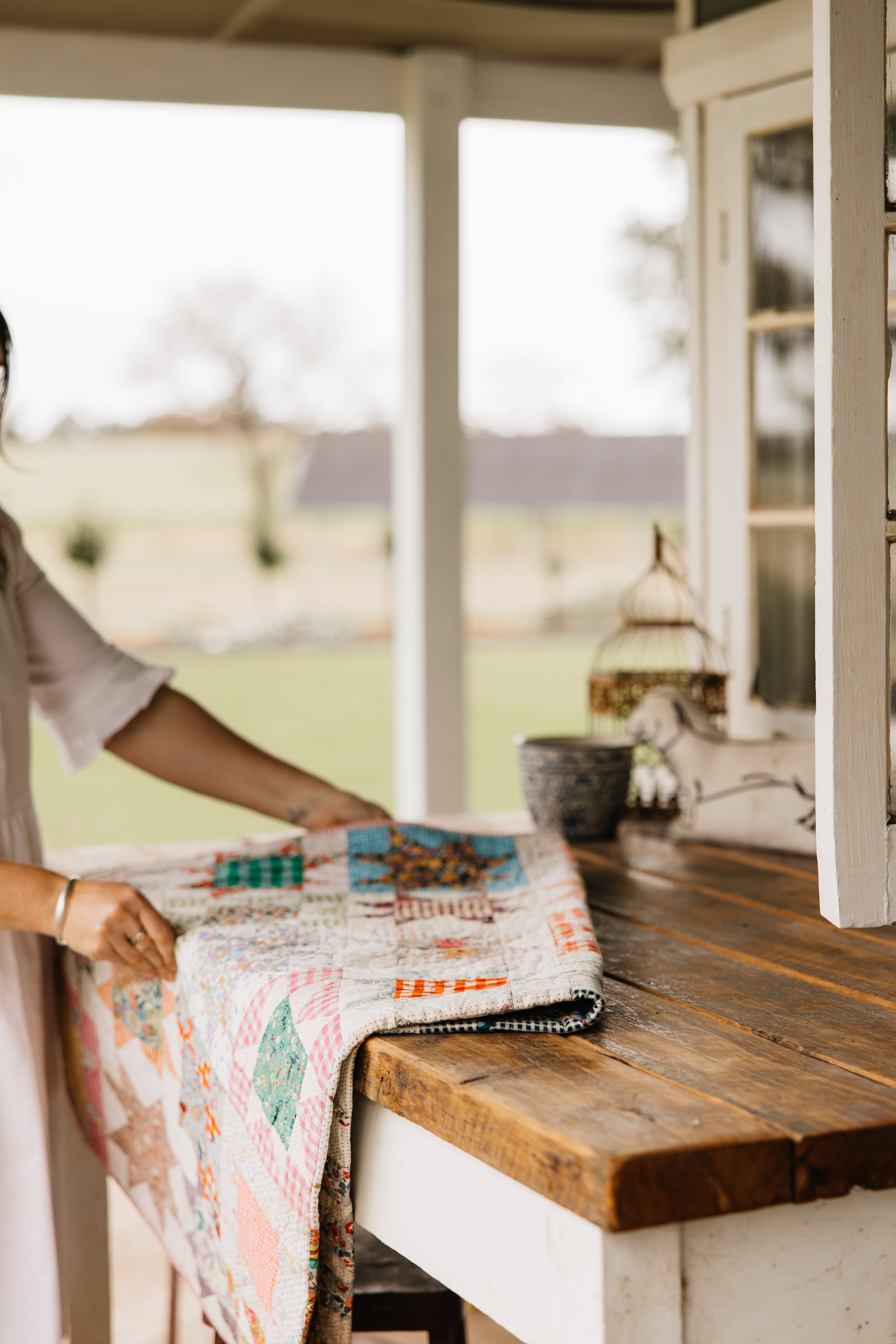 Person folding a colorful quilt on a rustic wooden table outdoors with a blurry background of trees and grass.