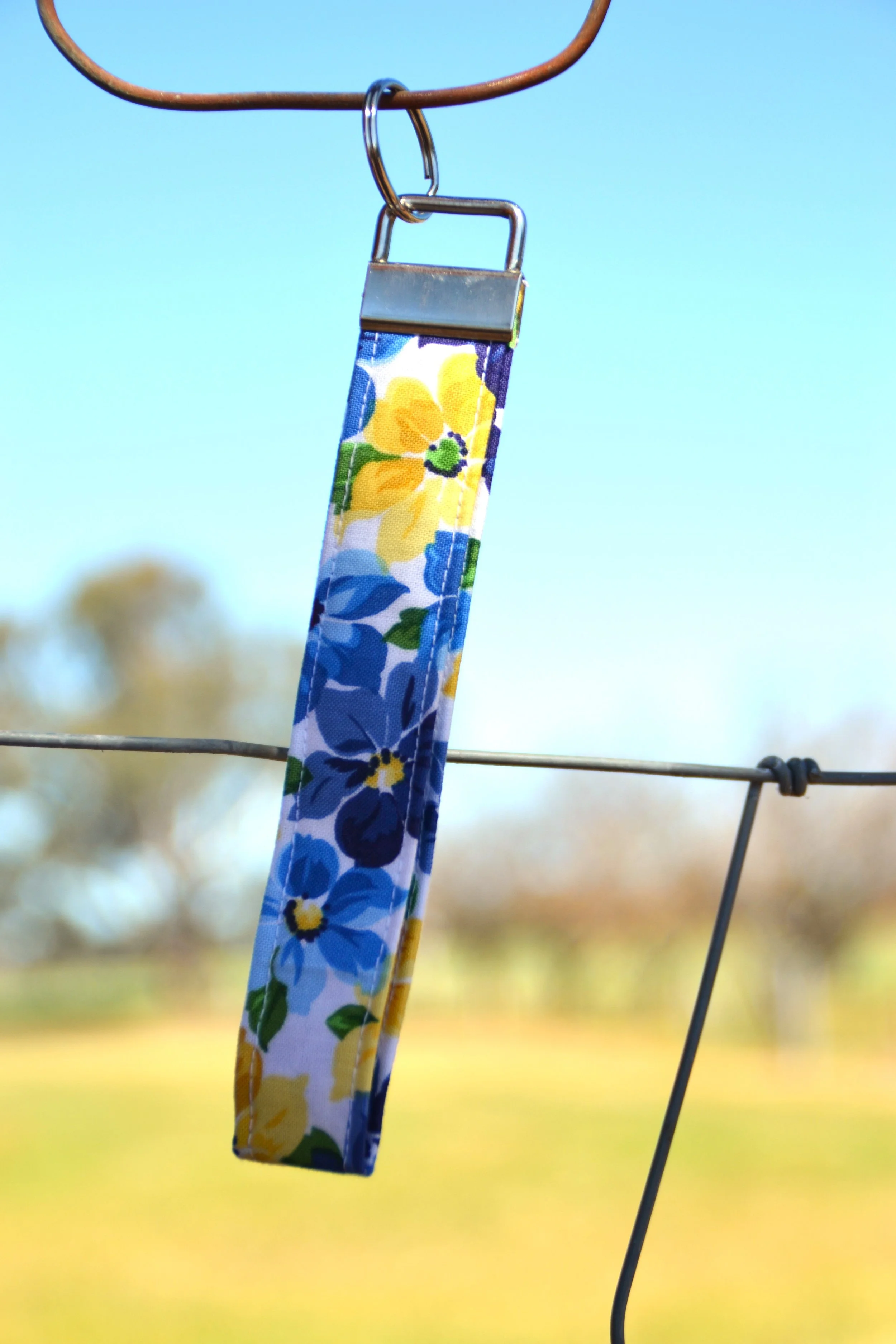 A fabric strap with a colorful floral pattern hanging from a metal hook on a wire fence with a blurred outdoor background.