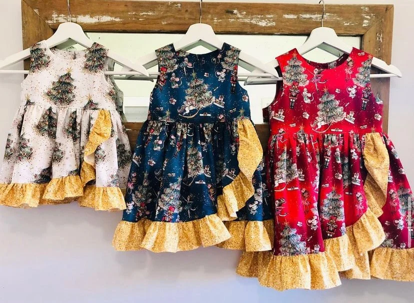 Three Christmas-themed dresses hanging on white hangers, each with a holiday print and yellow ruffle hem, displayed against a wooden frame background.