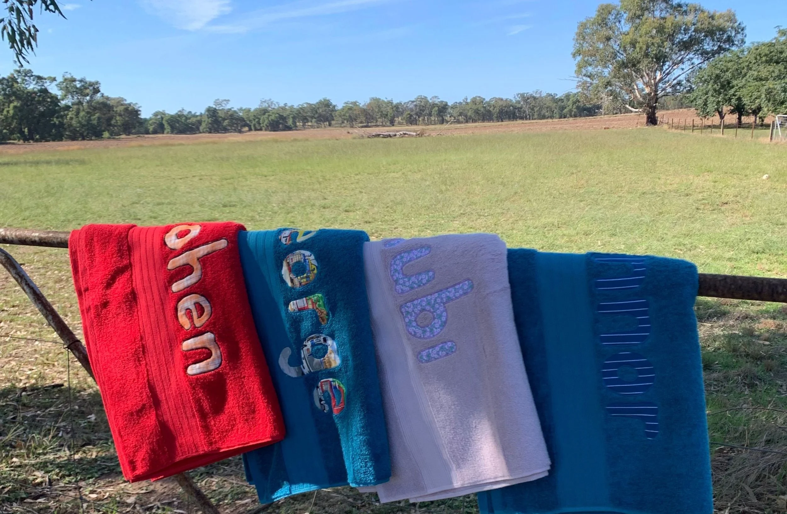 Four beach towels hanging on a rusty wire fence with a grassy field, trees in the distance, and a clear blue sky in the background.