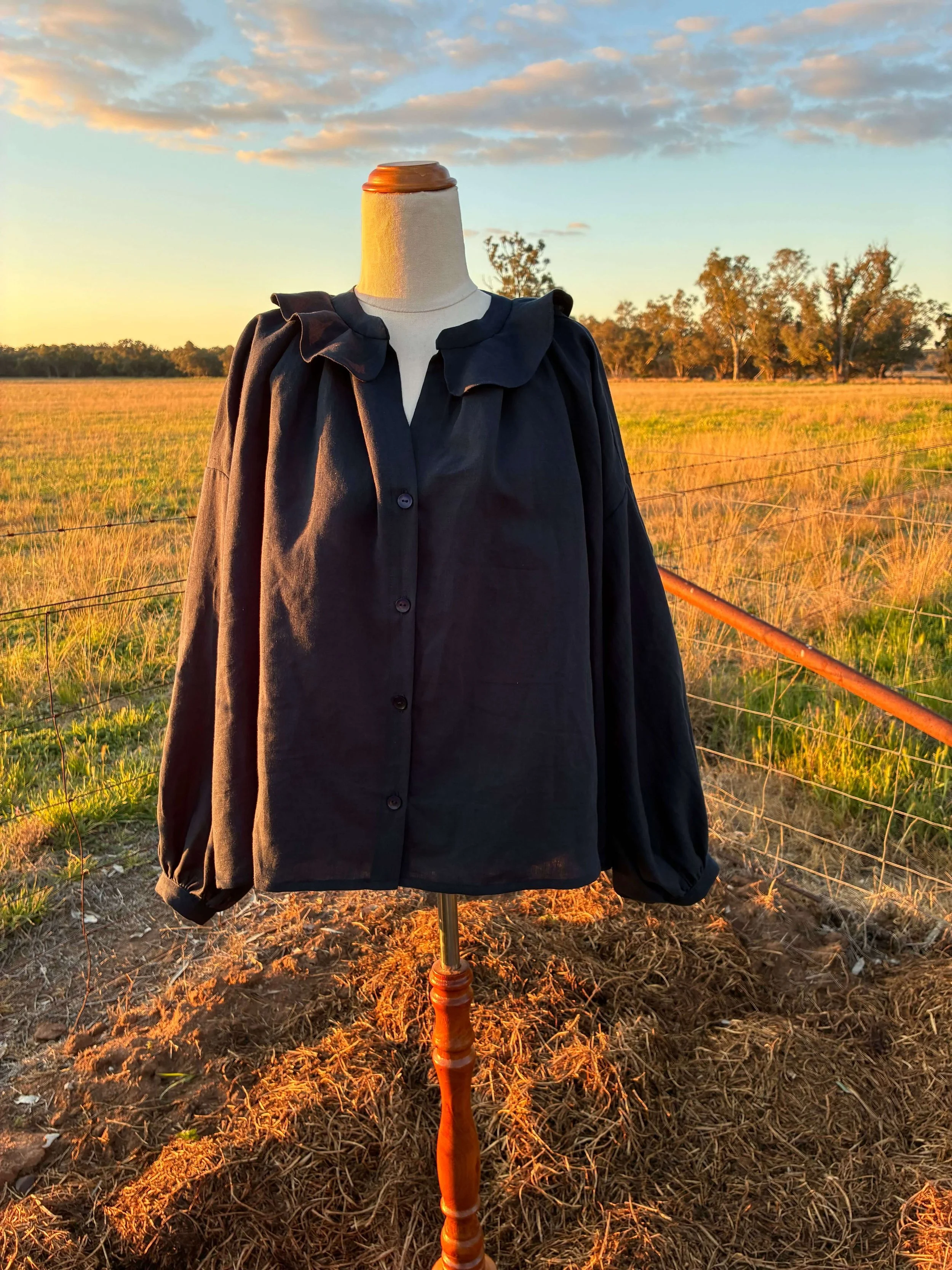 A mannequin dressed in a black jacket with a ruffled collar, positioned outdoors in a field at sunset.