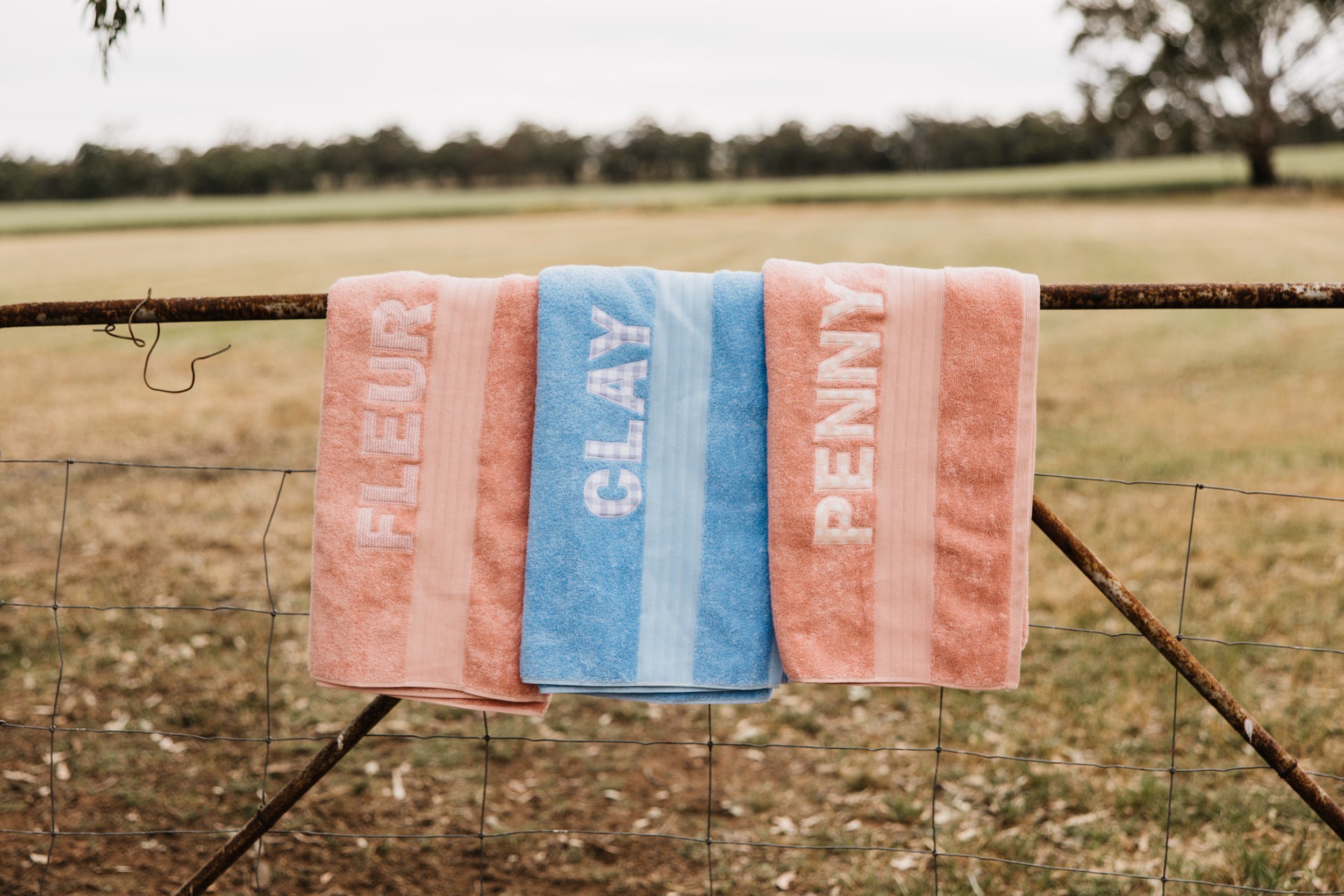 Three towels hanging on a rusty metal fence in a rural outdoor setting, with fields and trees in the background. The towels have the words "FLEUR," "CALAY," and "PENNY" embroidered on them.