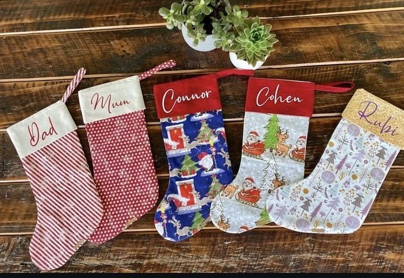 Five decorated Christmas stockings hanging on string, labeled for Dad, Mum, Connor, Cheen, and Ruba, with a plant in a white pot on wooden surface.