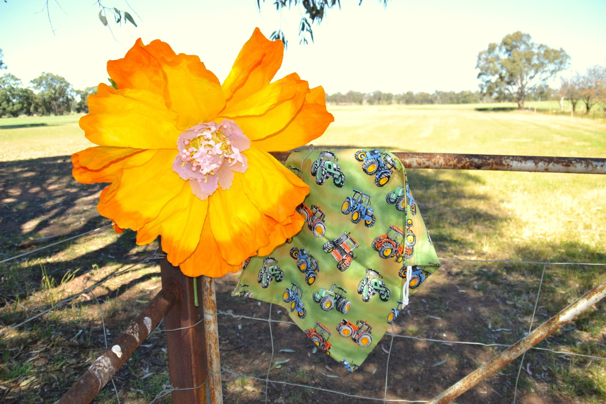 Large yellow and orange flower with pink center attached to a rusty metal fence. Cloth with tractor pattern draped from the flower on the fence in an open outdoor setting.