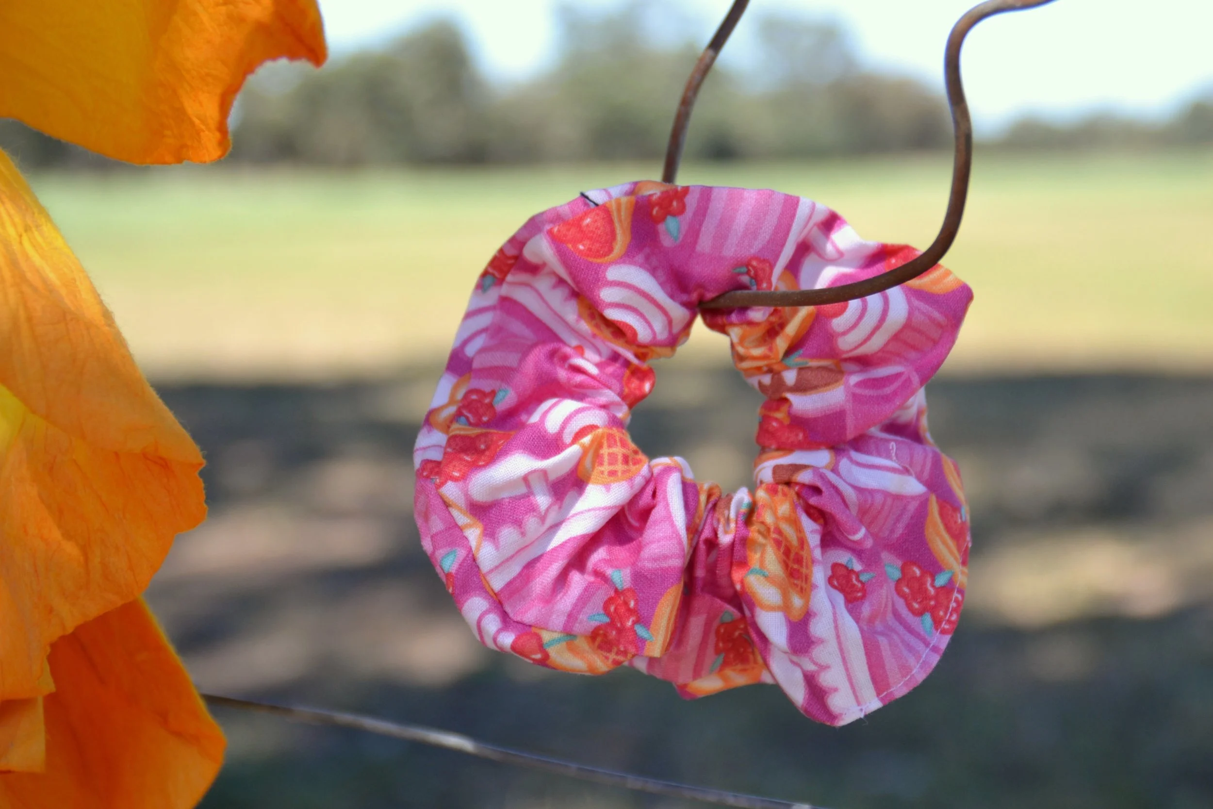 A pink fabric scrunchie with a colorful fruit pattern, hanging on a metal wire outdoors with a blurred landscape in the background.