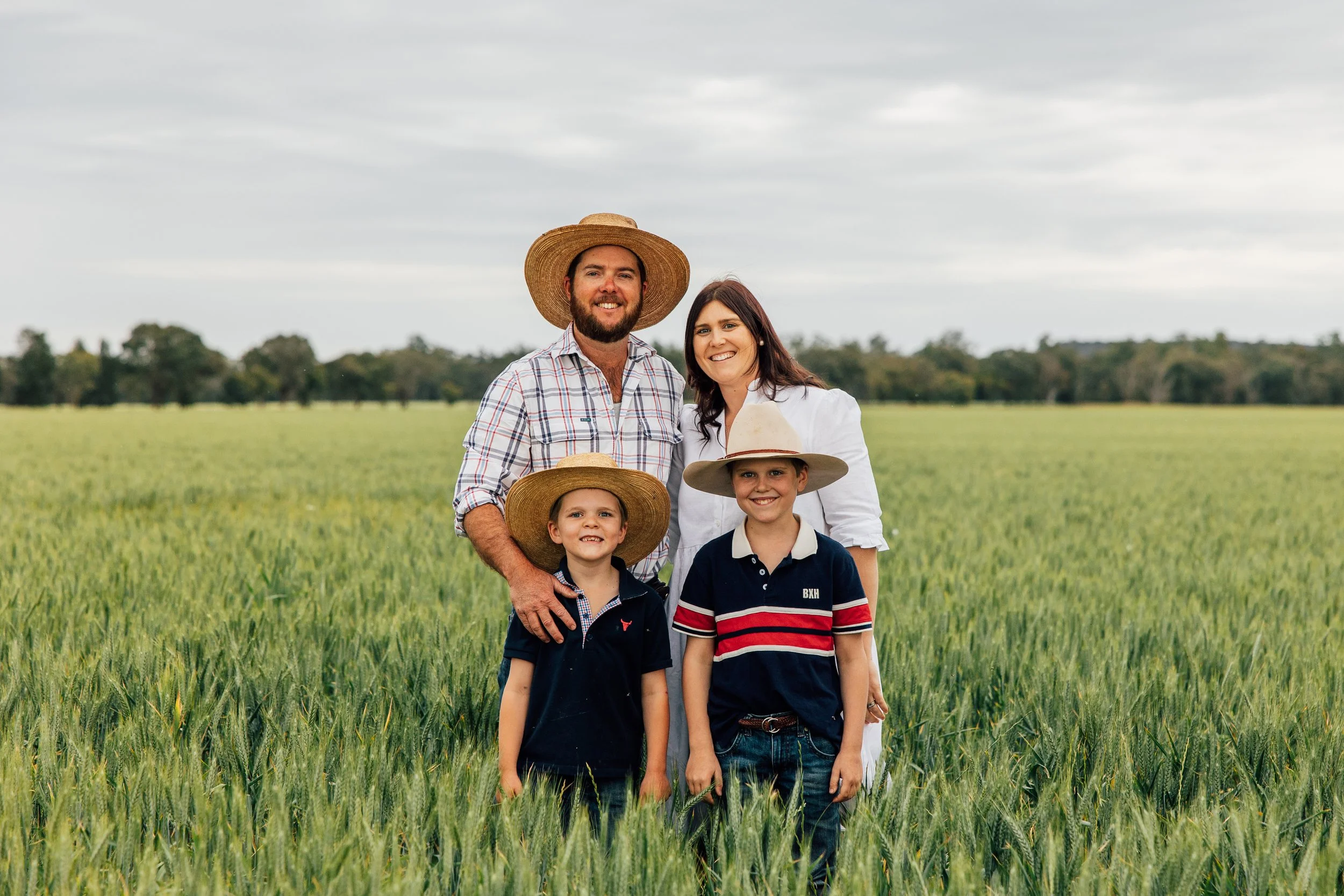 A family of four standing in a green field with trees and a cloudy sky in the background, all wearing hats and smiling.